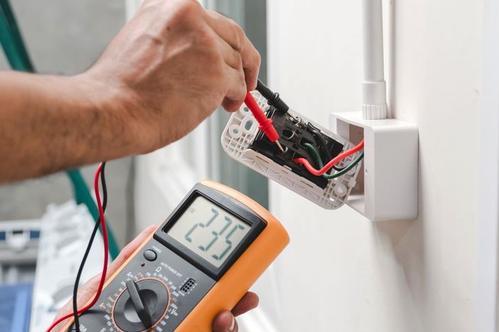 Technician testing exposed wall wiring with a multimeter in an electrical outlet box