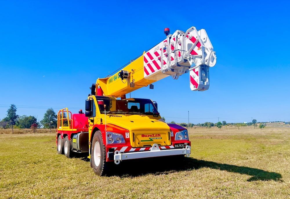 A Yellow And Red Crane Truck Is Parked In A Grassy Field — SureLift Crane Hire In Mackay, QLD