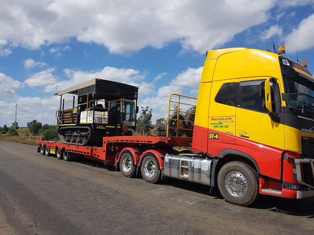 A Yellow And Red Semi Truck Is Carrying A Tractor Down A Road — SureLift Crane Hire In Nebo, QLD