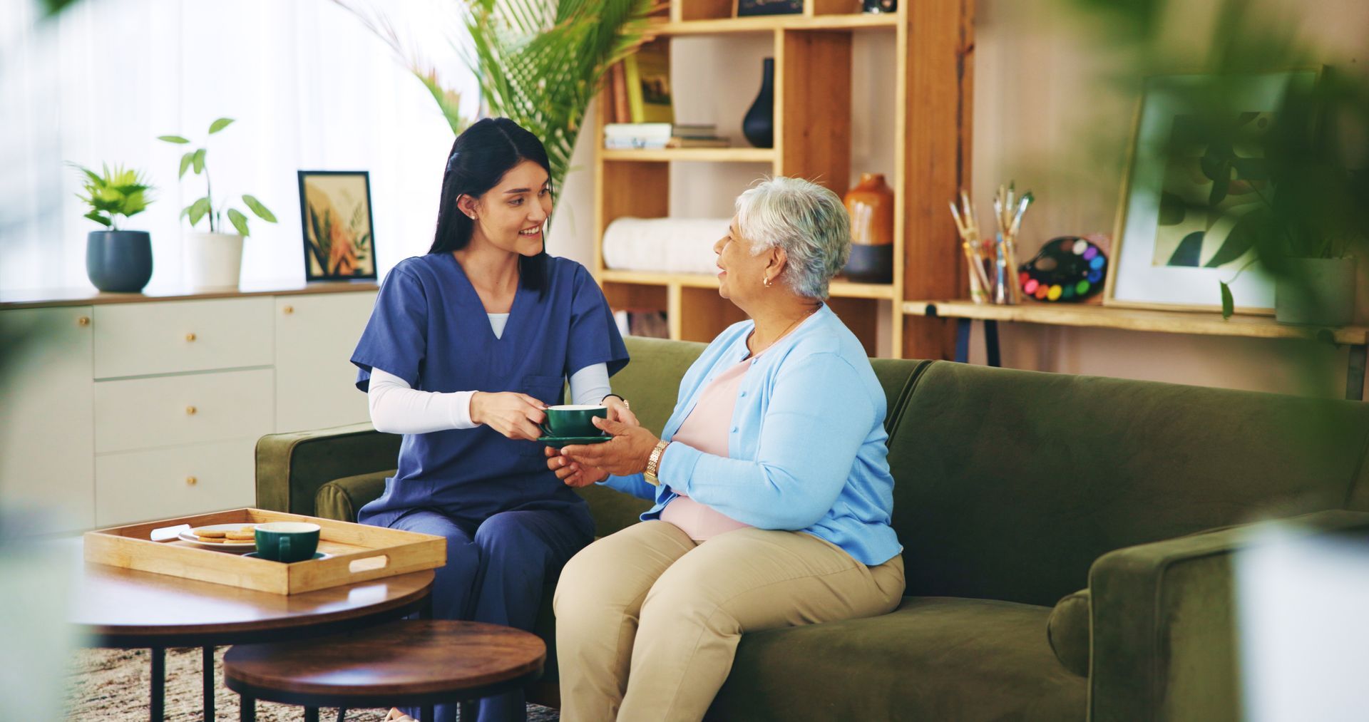 A caregiver in blue scrubs offers a green item to an older person seated on a green couch in a living room A caregiver in blue scrubs offers a green item to an older person seated on a green couch in a living room