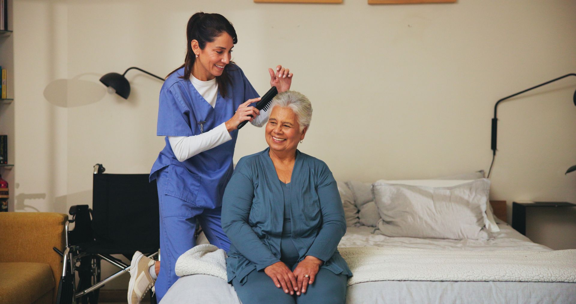 Caregiver combs the hair of an older adult woman sitting on a bed