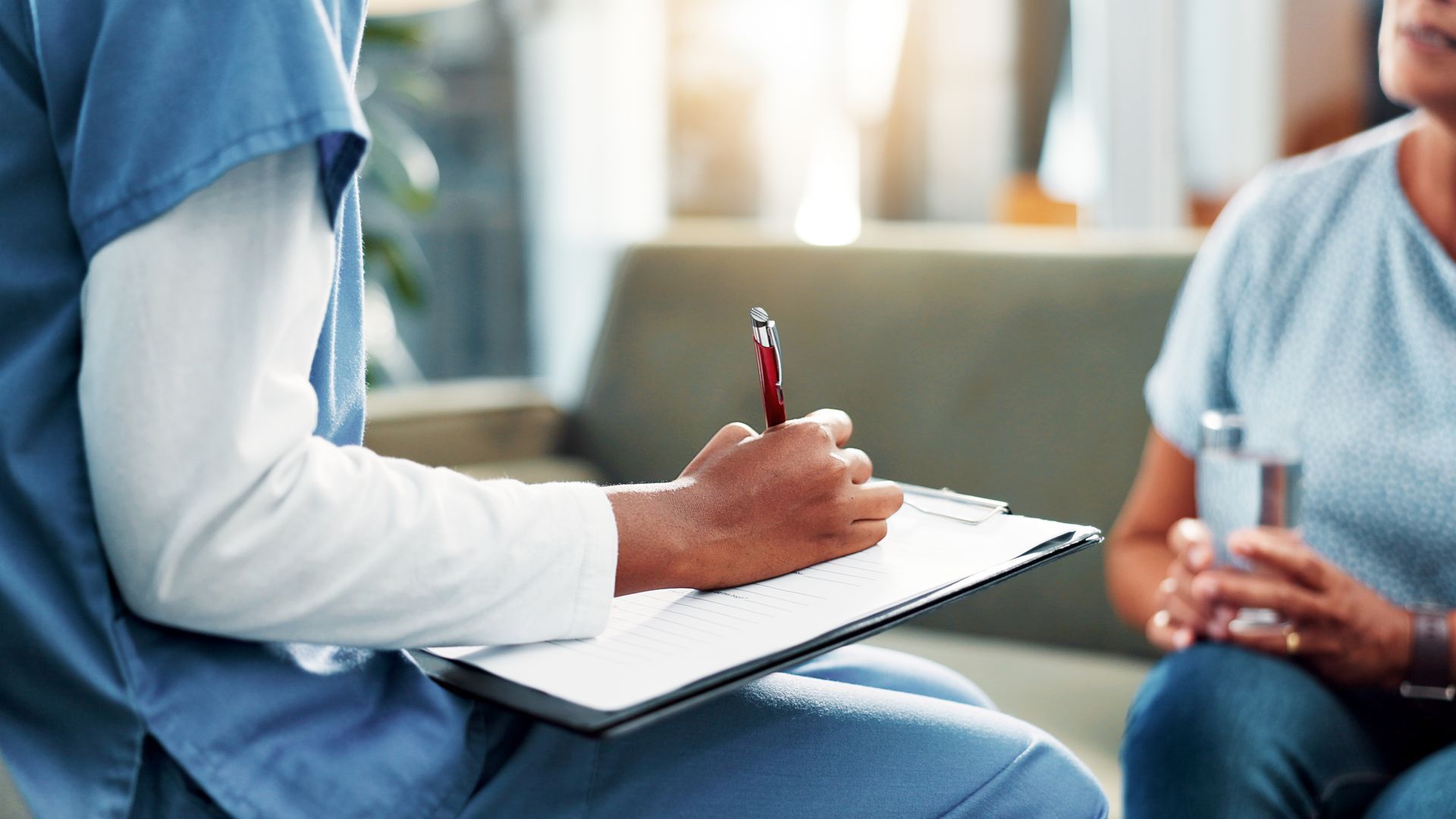 Person in scrubs writing on a clipboard, interviewing another person, in a home setting