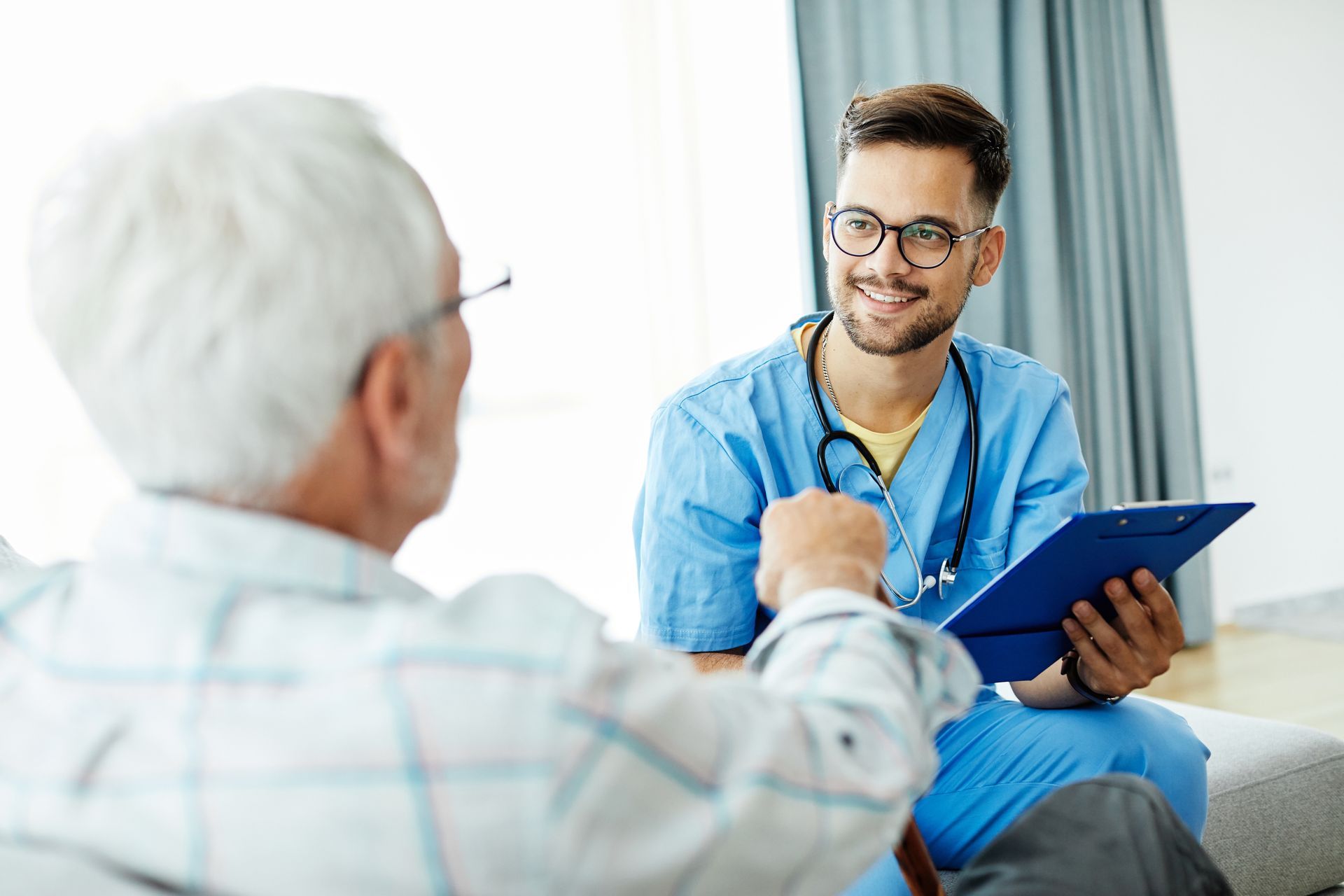 Healthcare worker in blue scrubs smiling while looking at elderly patient, holding clipboard