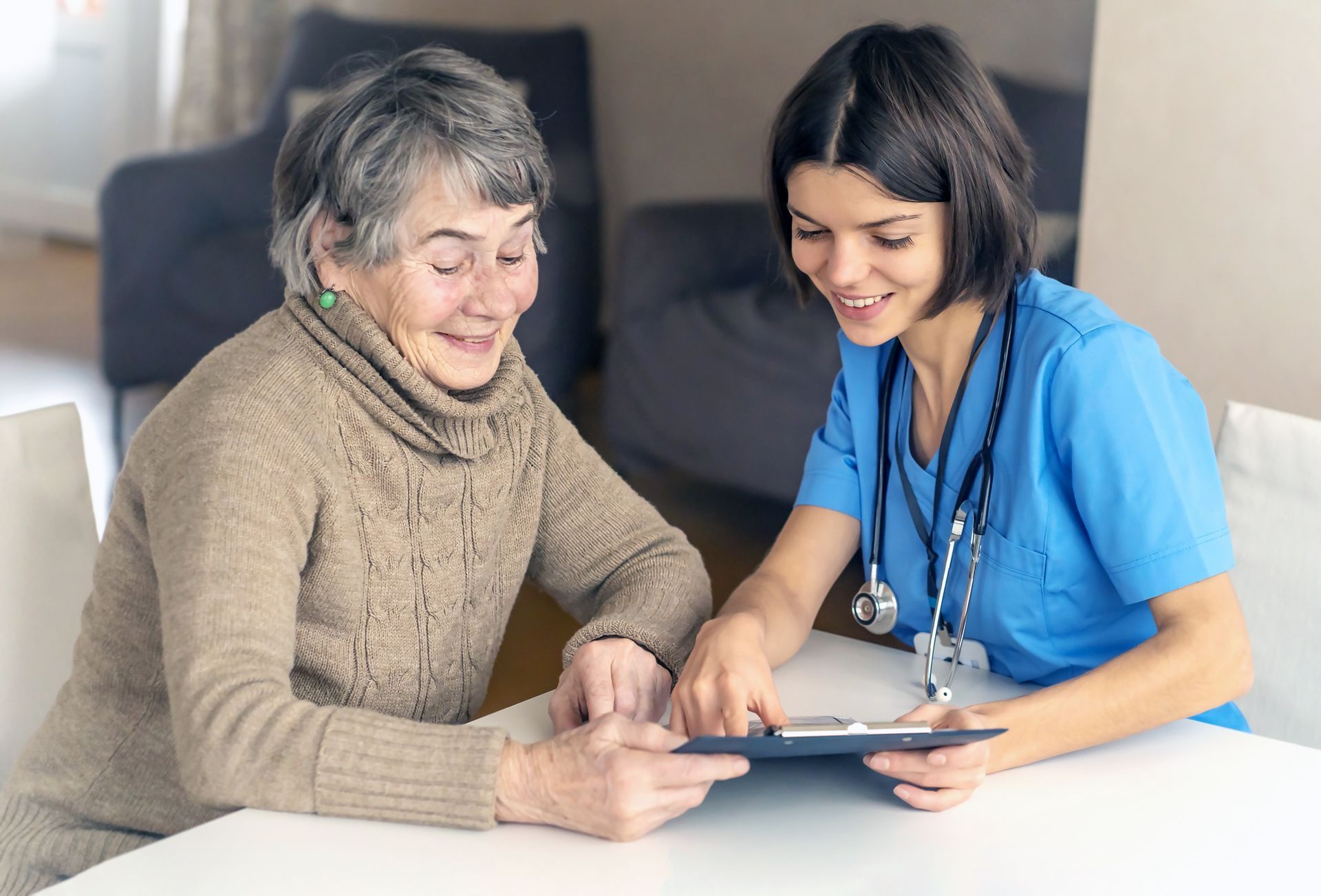 Woman and healthcare worker smiling, reviewing documents at a table. Blue scrubs, stethoscope visible Woman and healthcare worker smiling, reviewing documents at a table. Blue scrubs, stethoscope visible