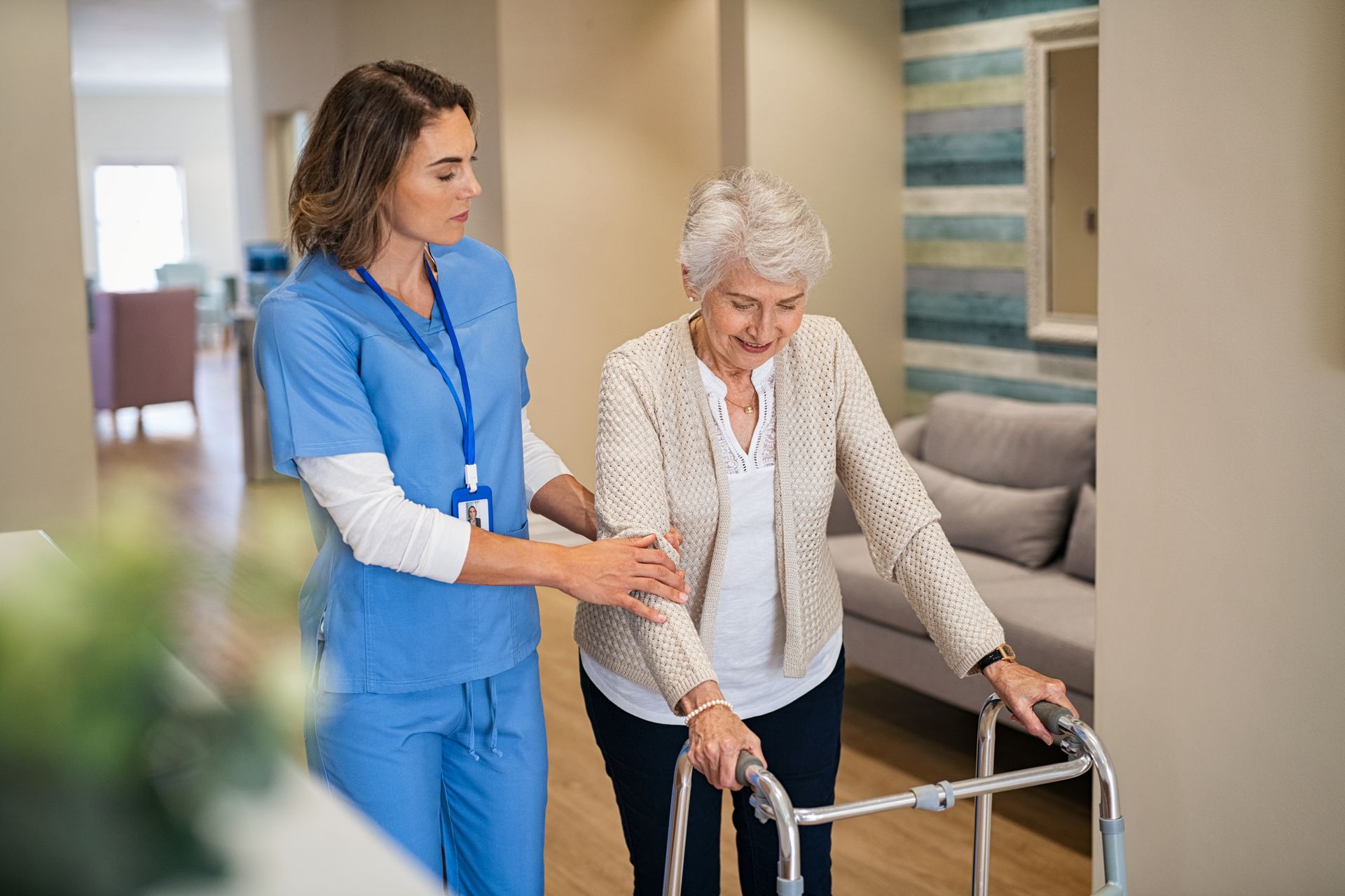 Nurse assisting a person using a walker in a hallway. Both are smiling Nurse assisting a person using a walker in a hallway. Both are smiling