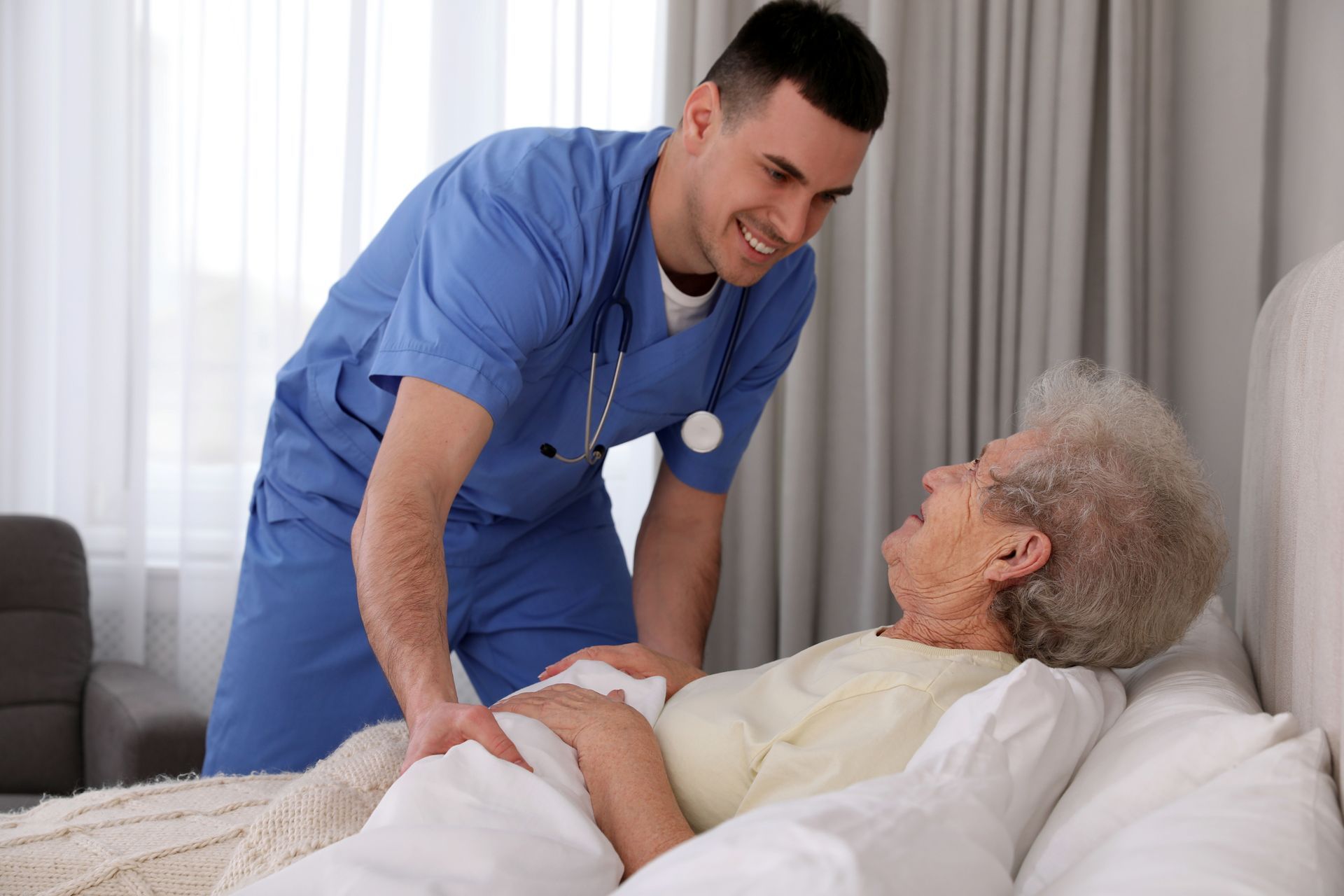 Healthcare worker in blue scrubs assisting an elderly person in bed, smiling and looking down Healthcare worker in blue scrubs assisting an elderly person in bed, smiling and looking down