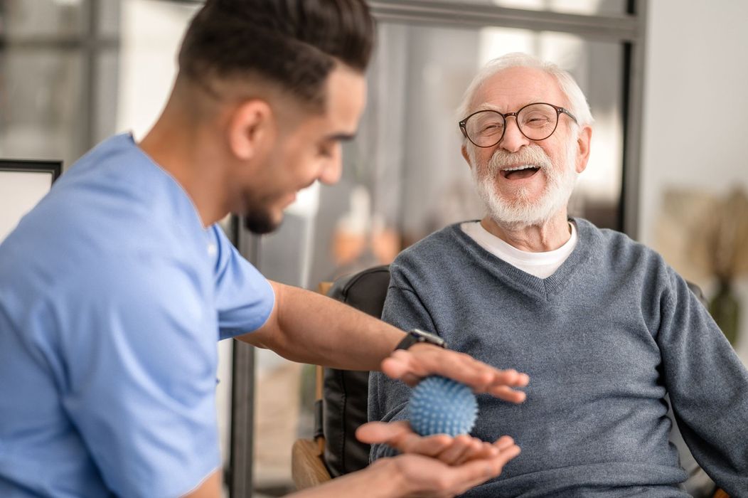Caregiver assists elderly man with a therapy ball. Both smiling in an indoor setting