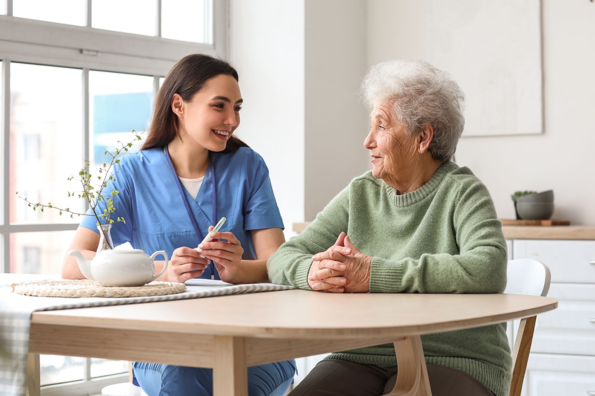 A healthcare worker in blue scrubs talking with an older person seated at a table