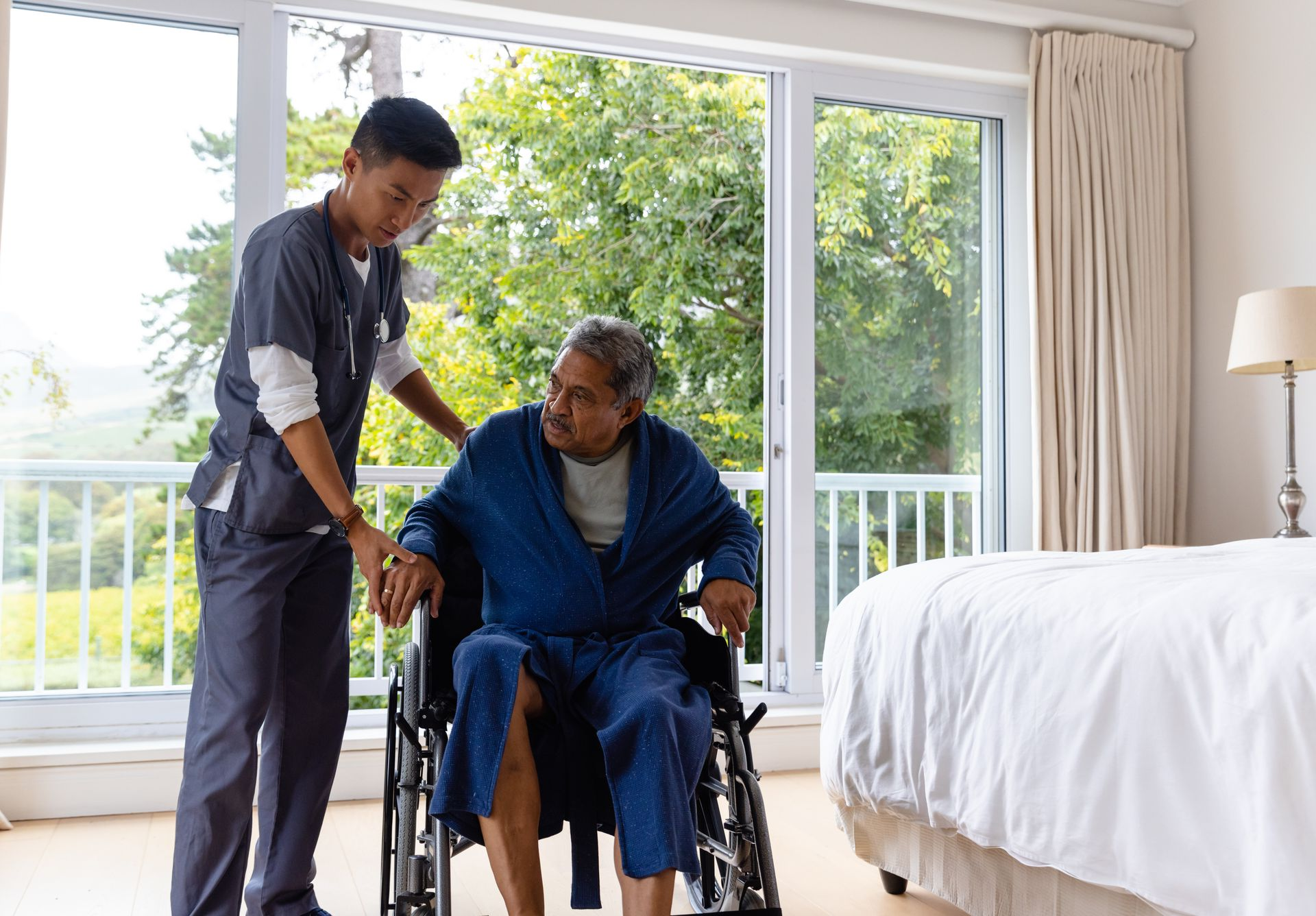 Caregiver assisting a person using a walker in a room; the caregiver wears blue scrubs
