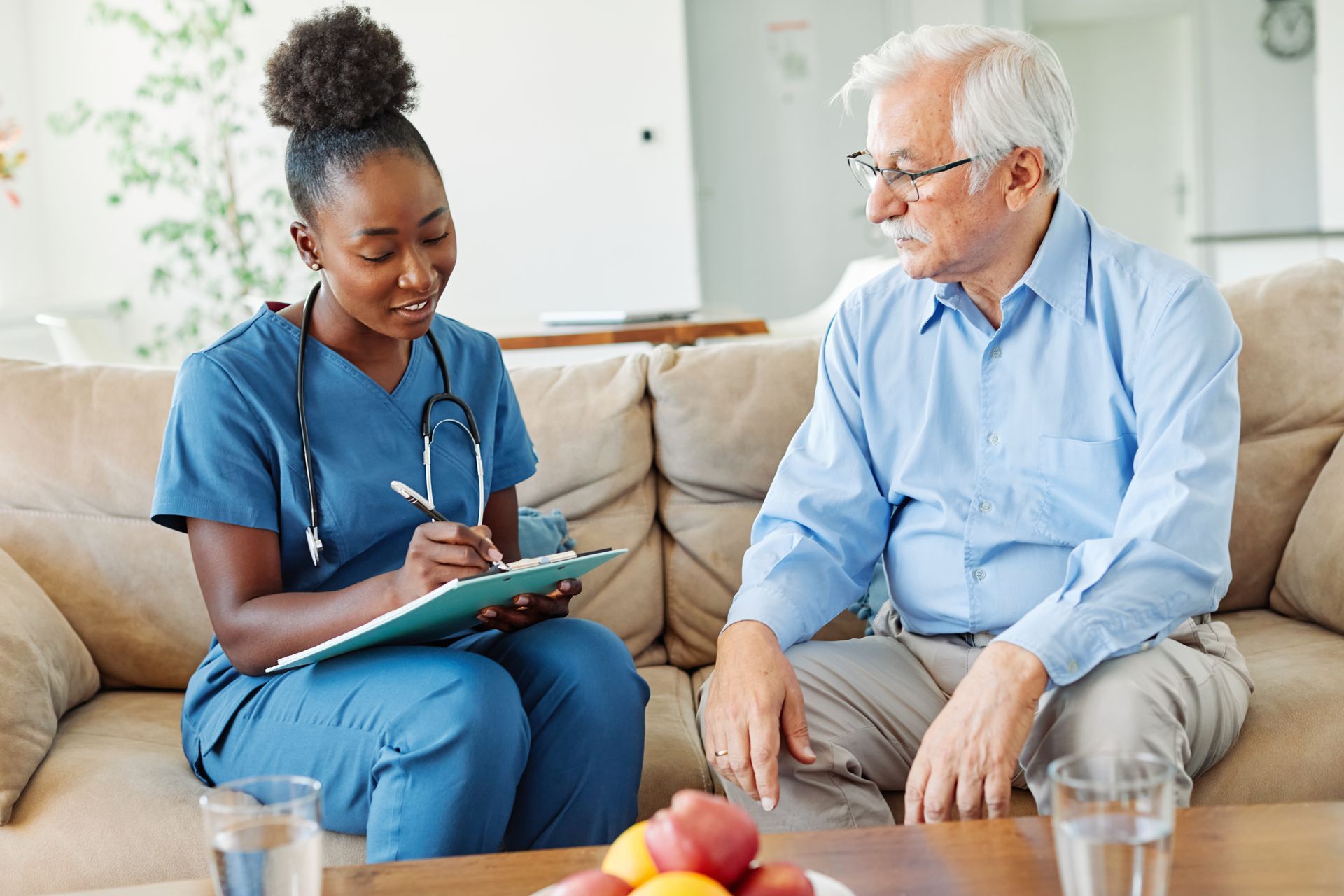 Nurse in blue scrubs, writing on a clipboard while speaking with an older person on a sofa Nurse in blue scrubs, writing on a clipboard while speaking with an older person on a sofa