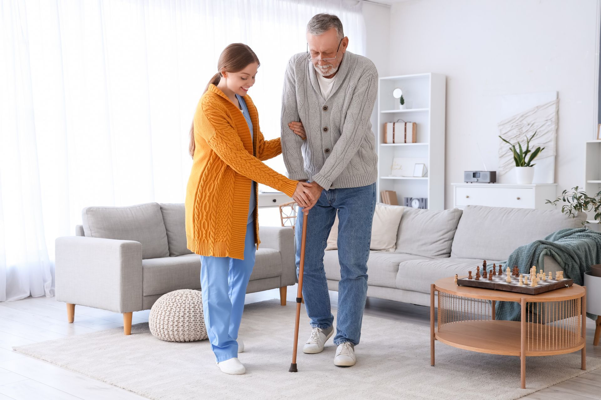 Woman in yellow cardigan helps man with cane walk in a living room