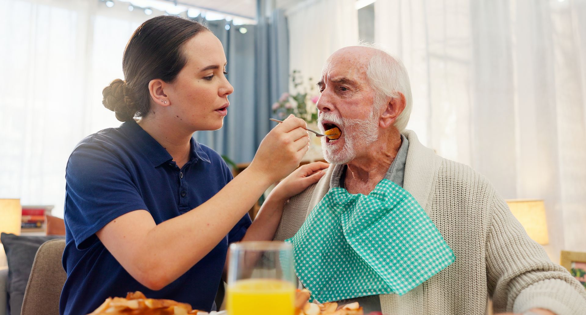 Caregiver feeding an elderly person with a bib in a dining room