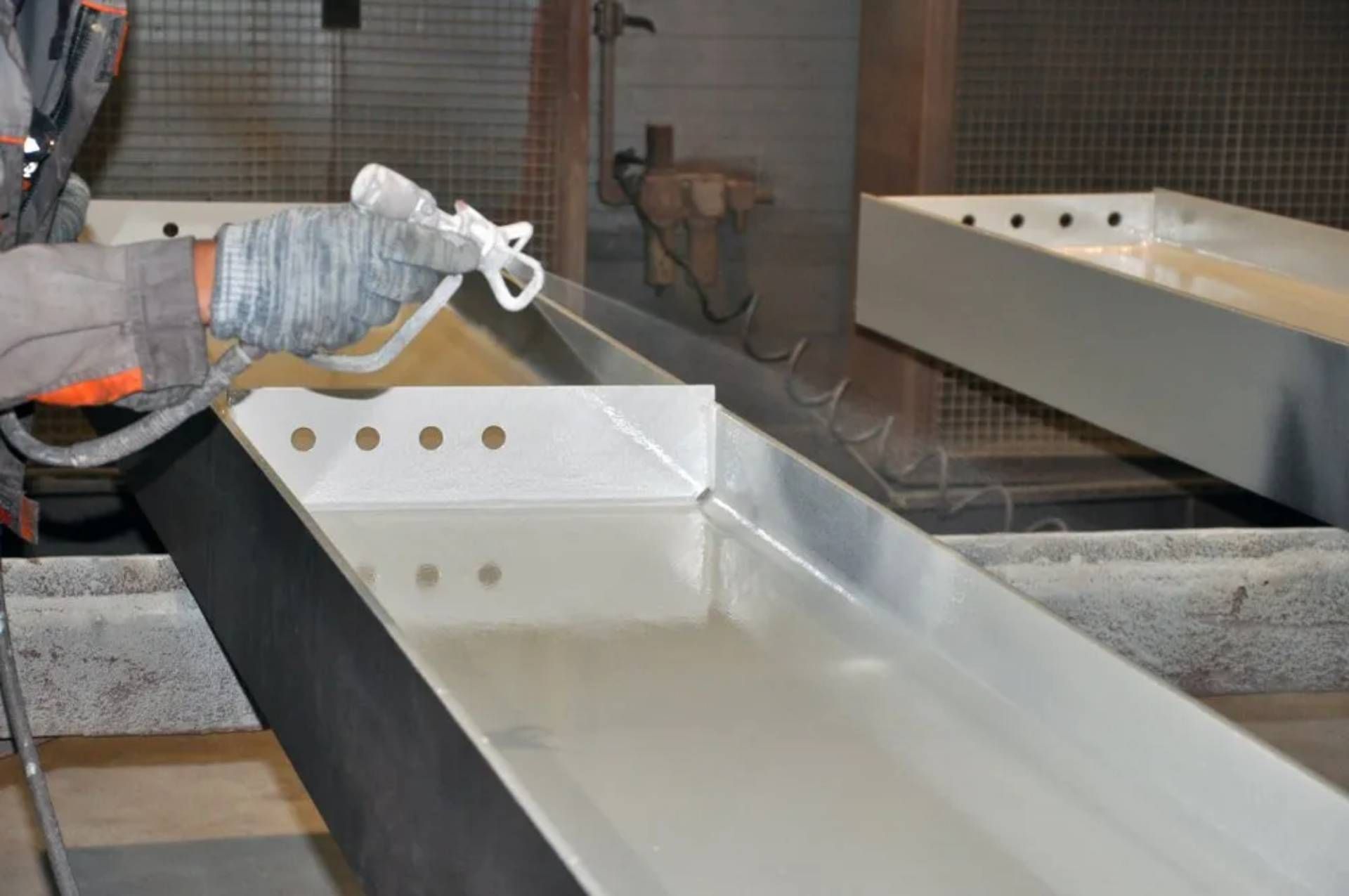 Persona che spruzza vernice bianca all'interno di una mangiatoia metallica. Un'altra mangiatoia è appesa lì vicino. Ambiente industriale.