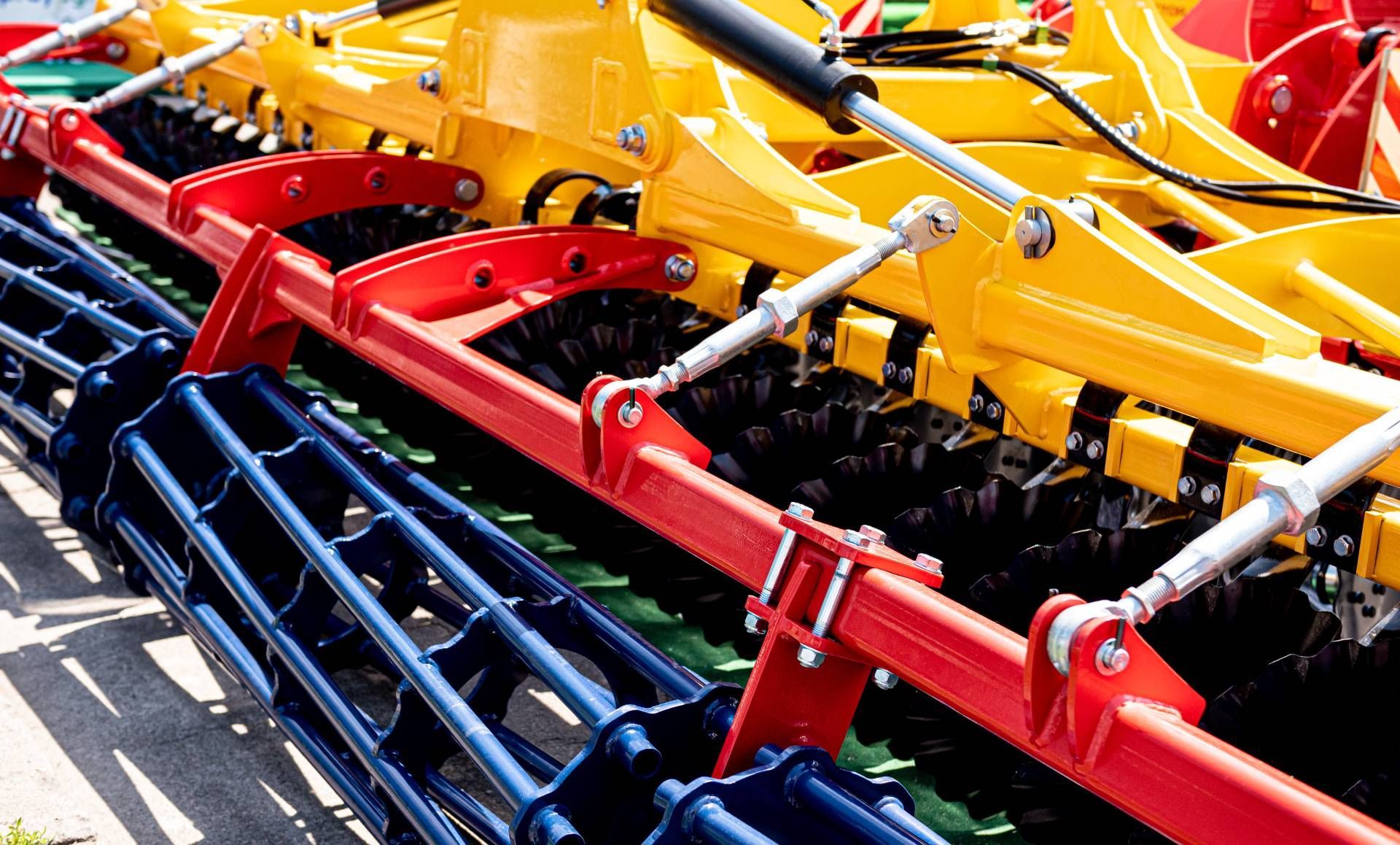 Primo piano di un attrezzo agricolo con componenti metallici rossi e gialli; sono visibili file di rebbi e rulli.