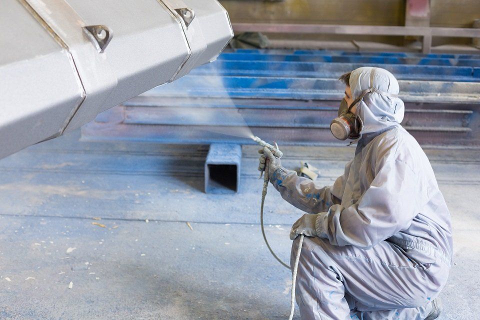 Persona con tuta protettiva che spruzza vernice su un oggetto metallico in un'officina.