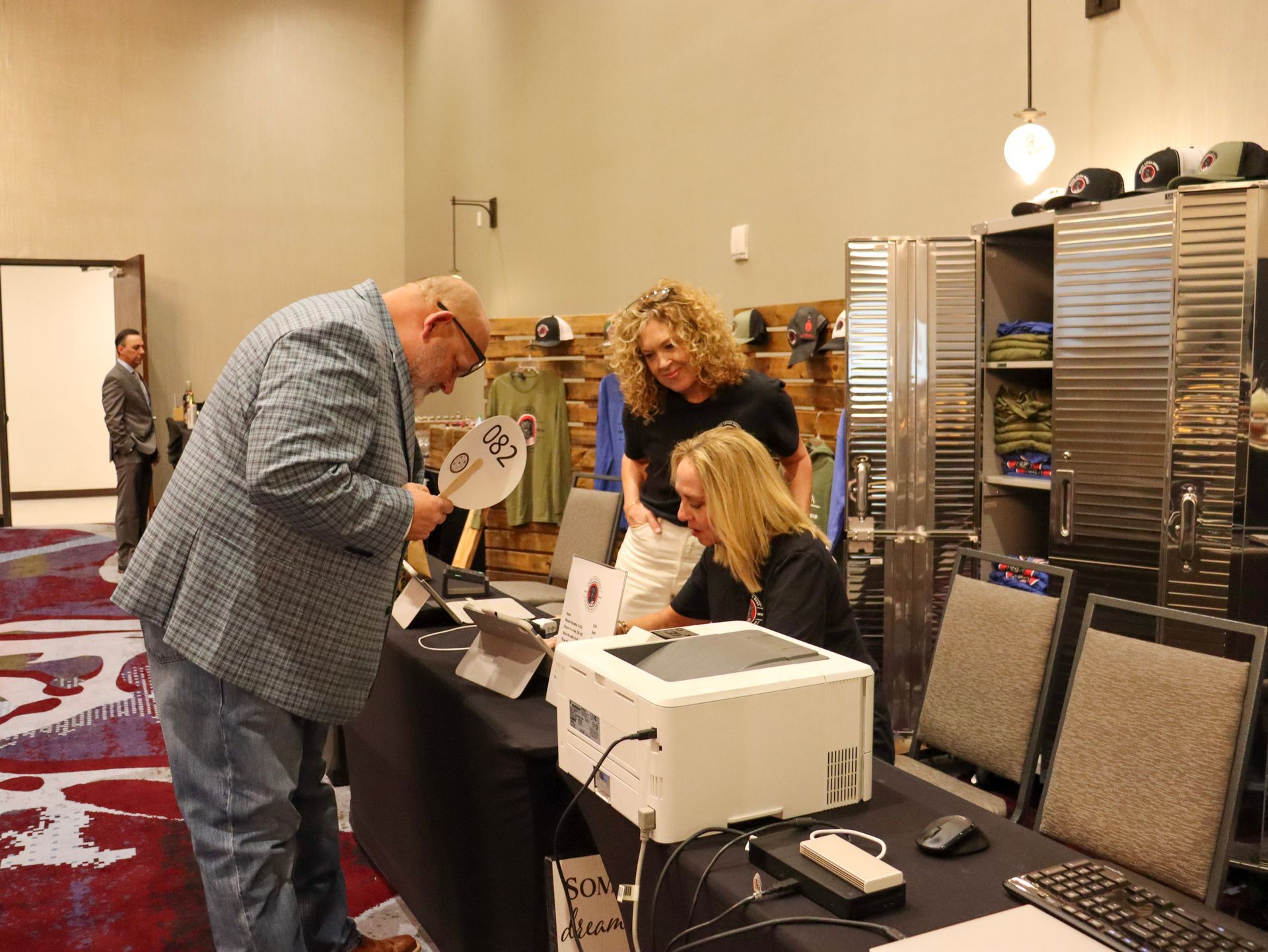 A person interacts with staff at a registration desk, holding a small sign while another staff member operates a printer.