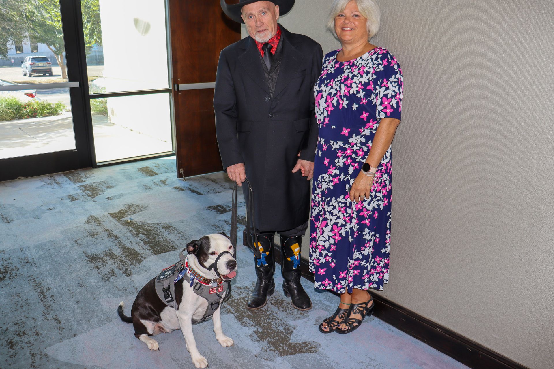 A man in a black suit and cowboy hat stands with a woman in a floral dress and a service dog in a building interior.