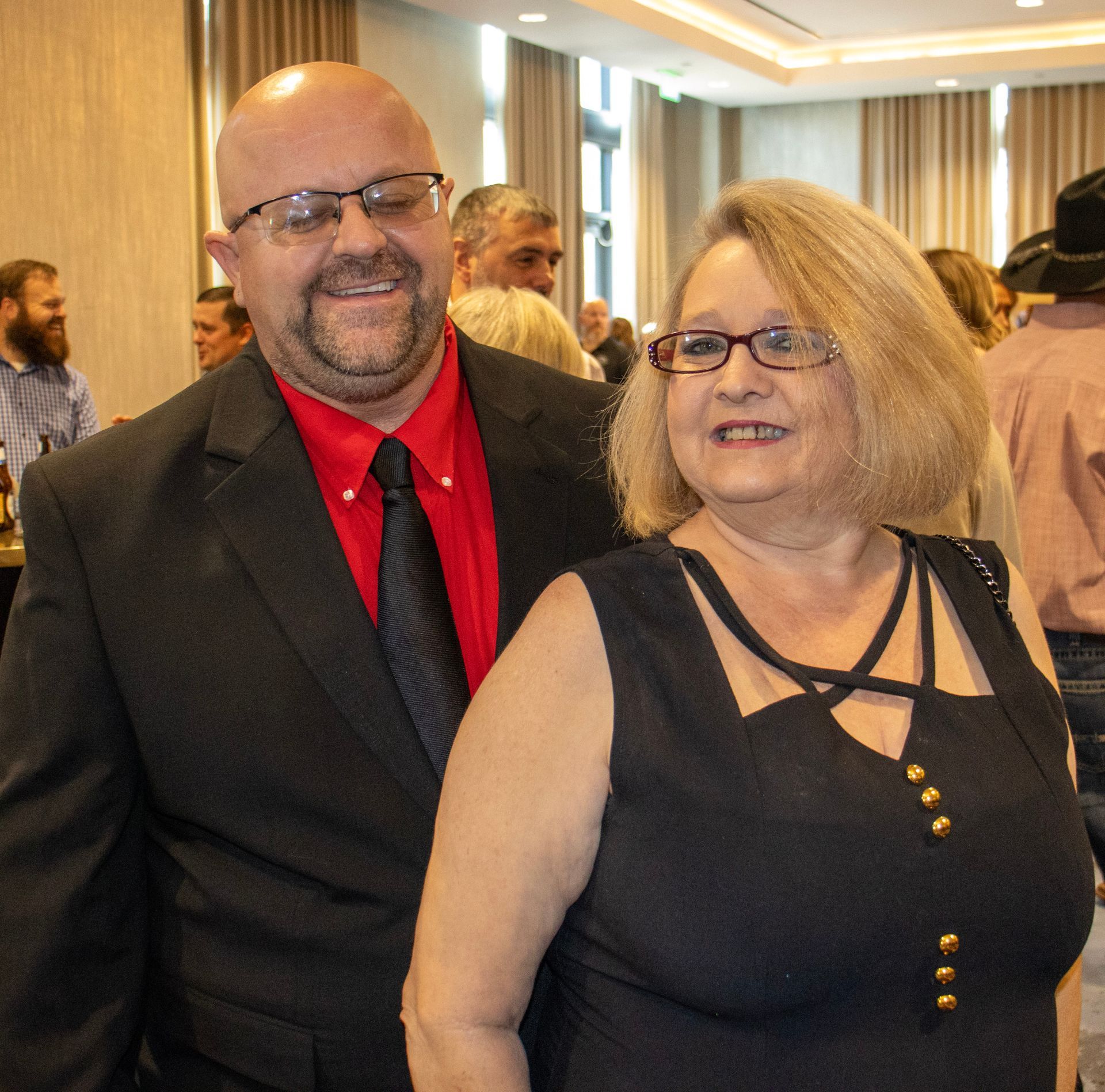 A couple stands close together at an indoor event, both smiling. The man wears a dark suit and red shirt; the woman, black.