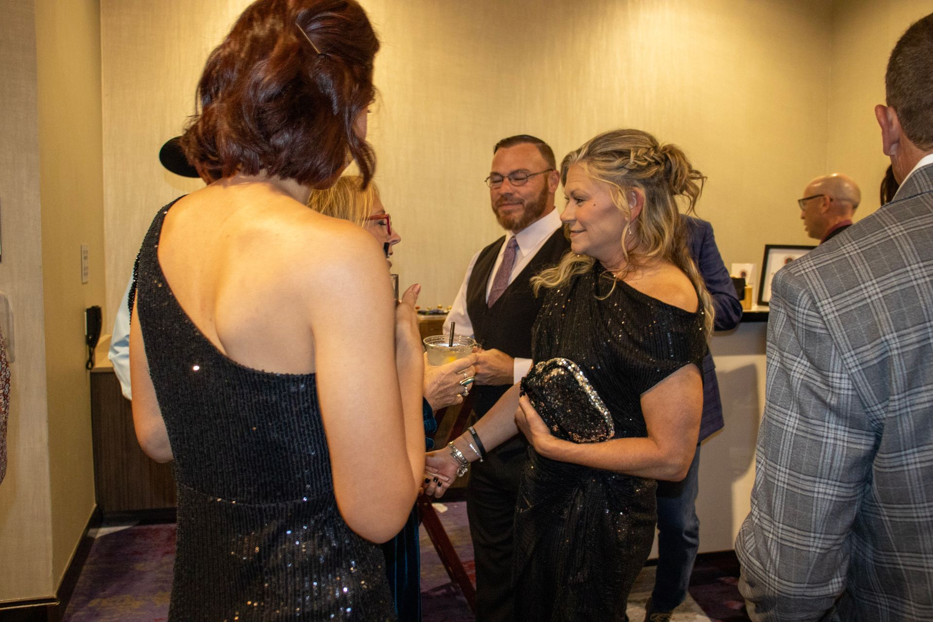 A group of people in formal attire stand in a room, socializing and holding drinks at an event.