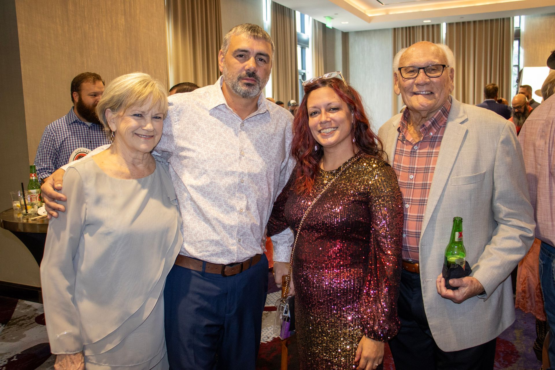 Four people stand posing together at an indoor event, with a man on the right holding a bottle.