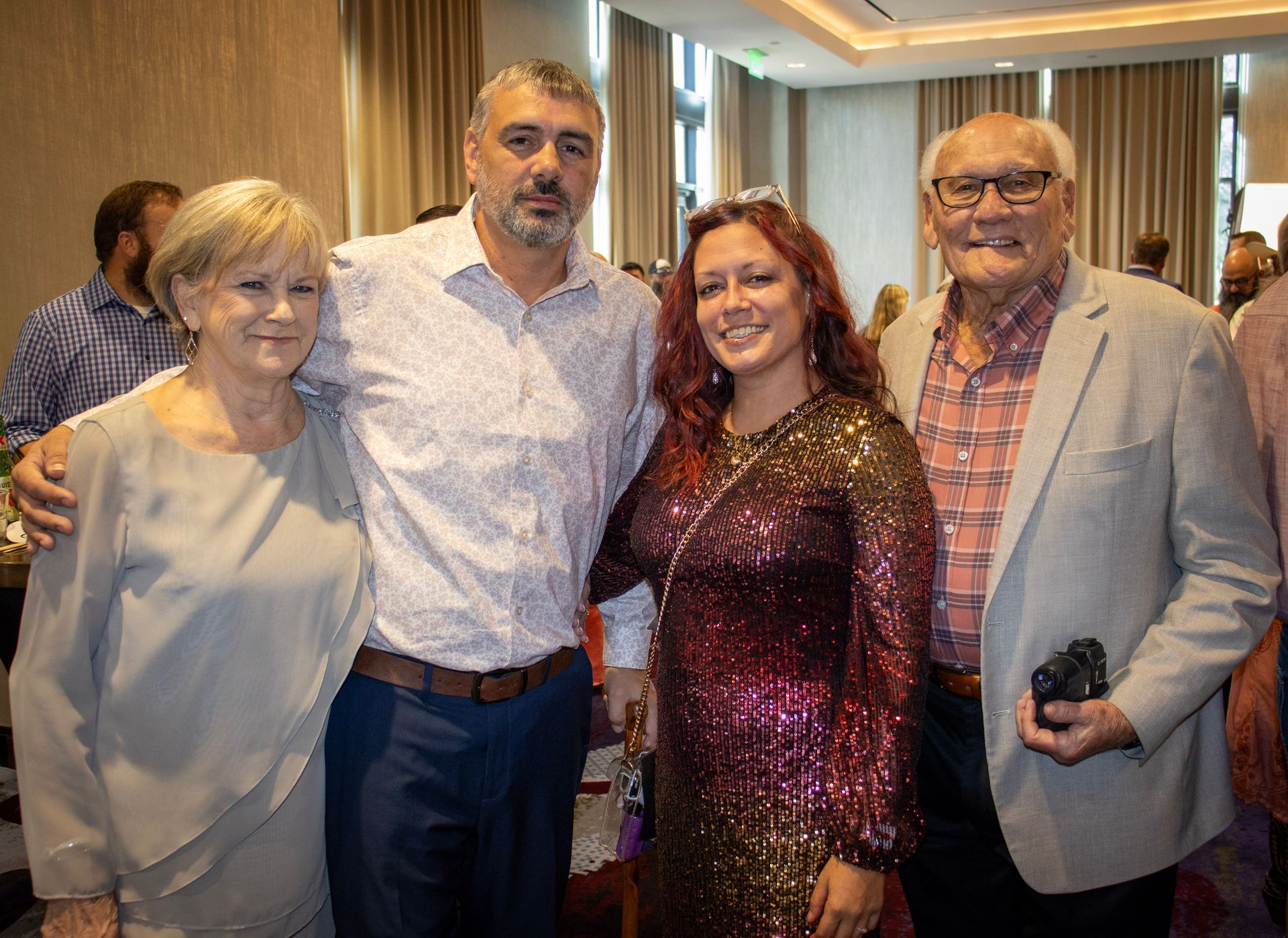 Four people pose for a photo indoors, wearing formal attire, with others blurred in the background.