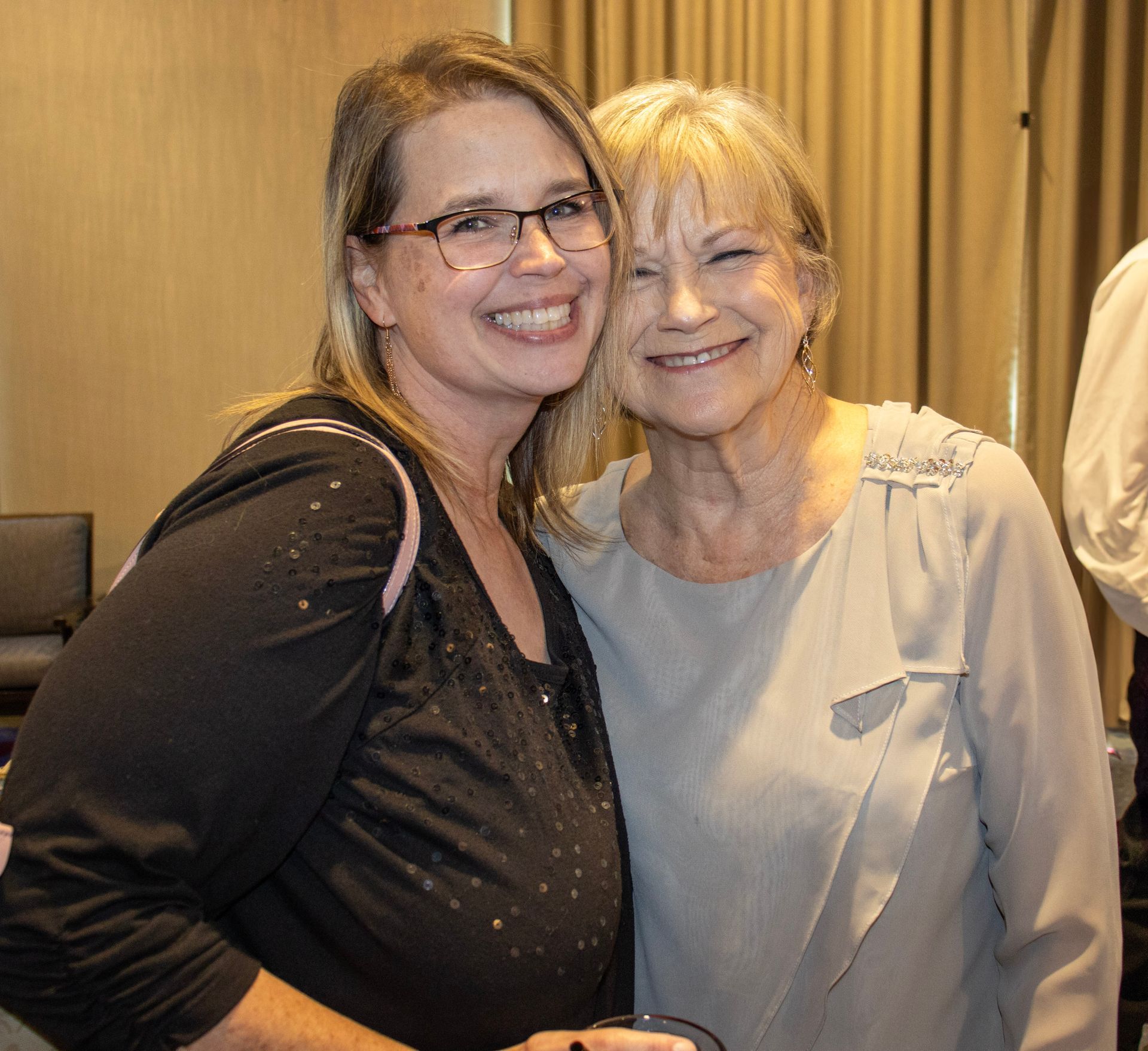 Two people smiling, standing side by side in an indoor setting with beige curtains in the background.