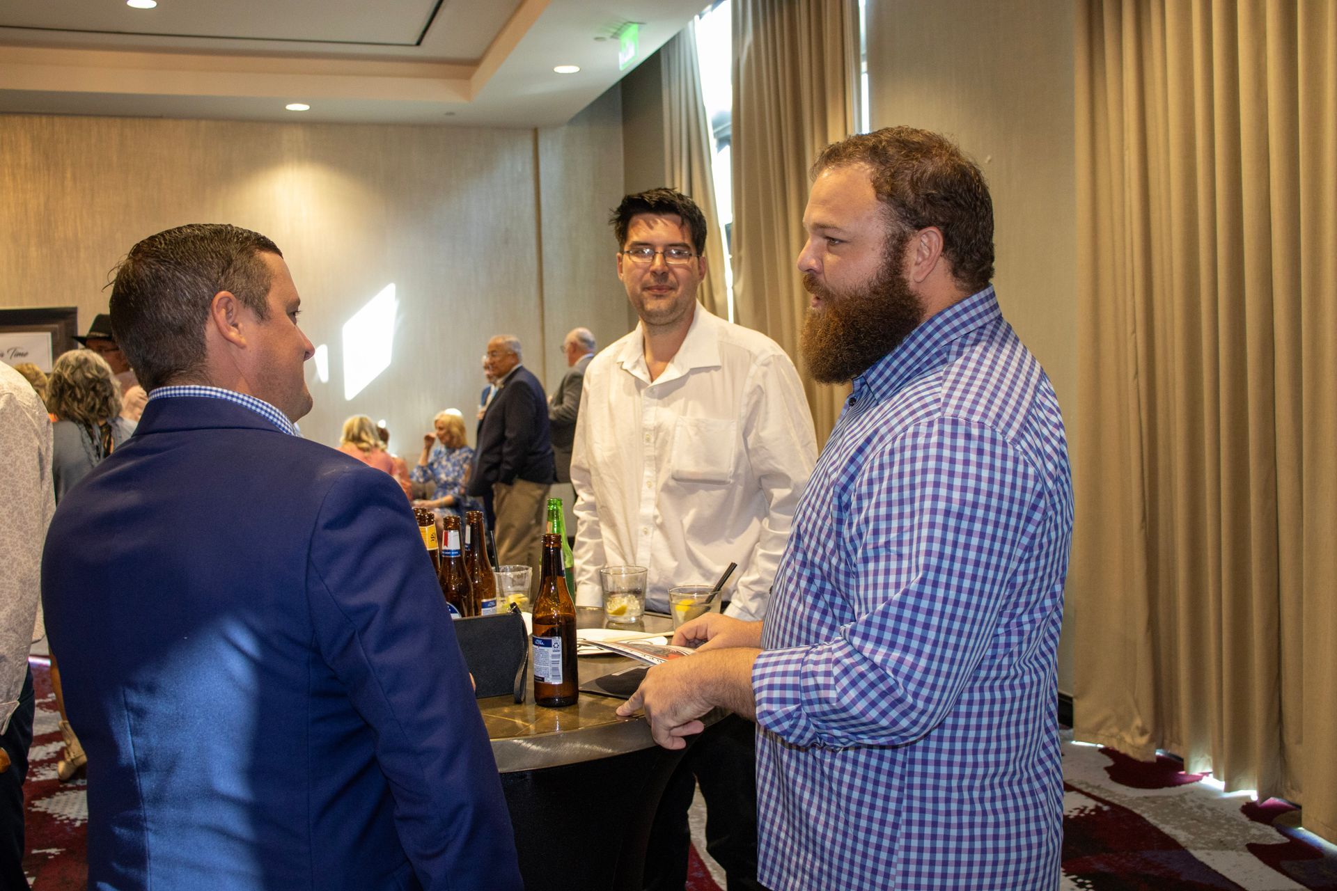 Three people converse at a bar table in a hotel conference room, with one person wearing a plaid shirt.
