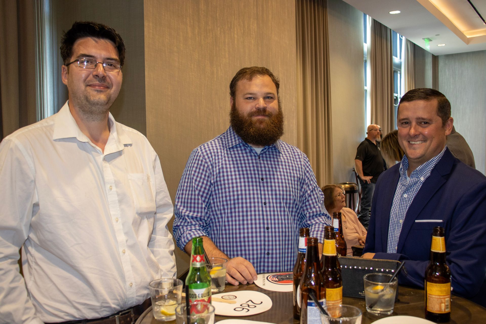 Three people standing around a table with drinks in a room with floor-to-ceiling curtains.