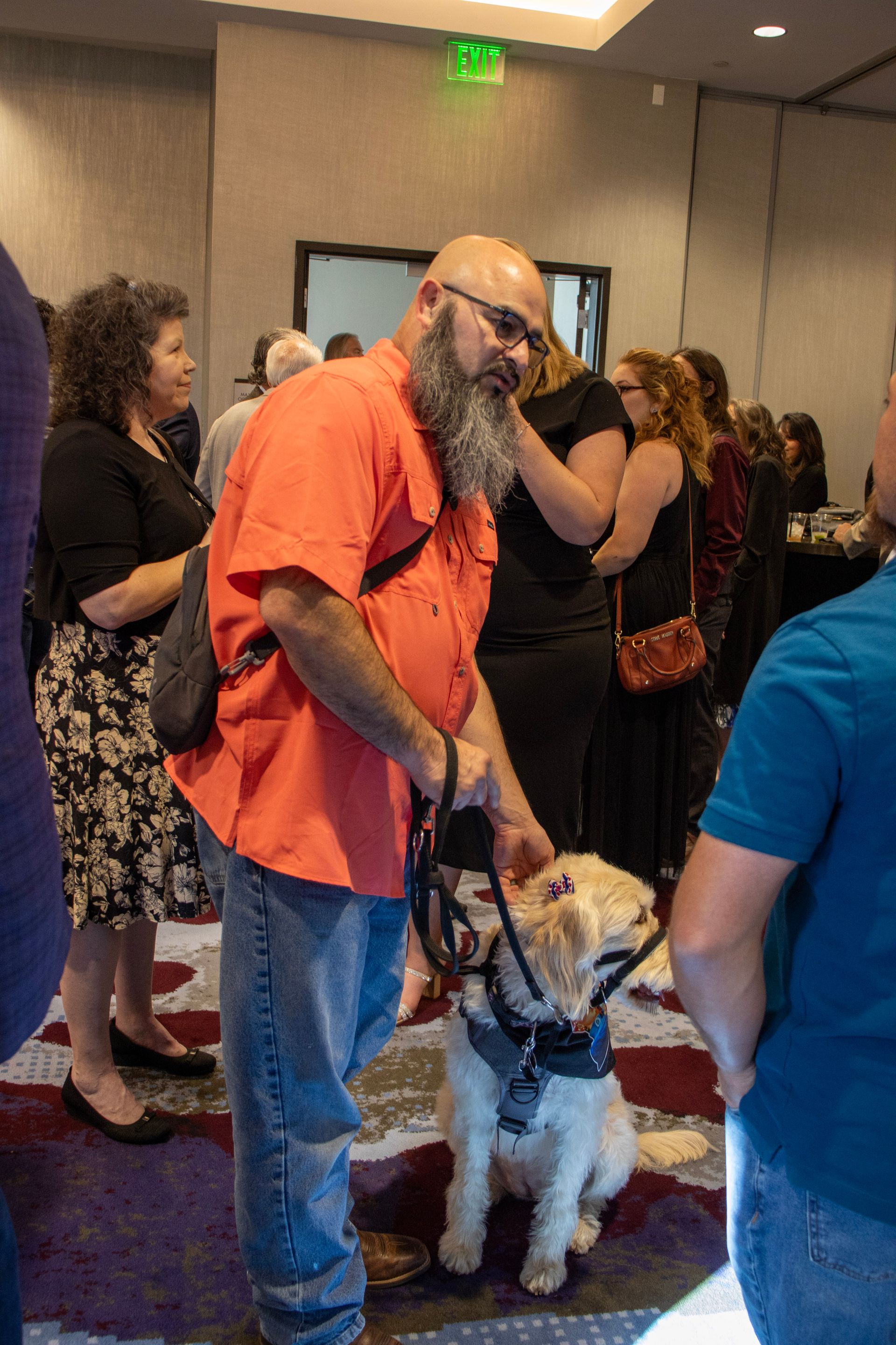 A person with a beard in an orange shirt interacts with their light-colored service dog in a busy conference room.