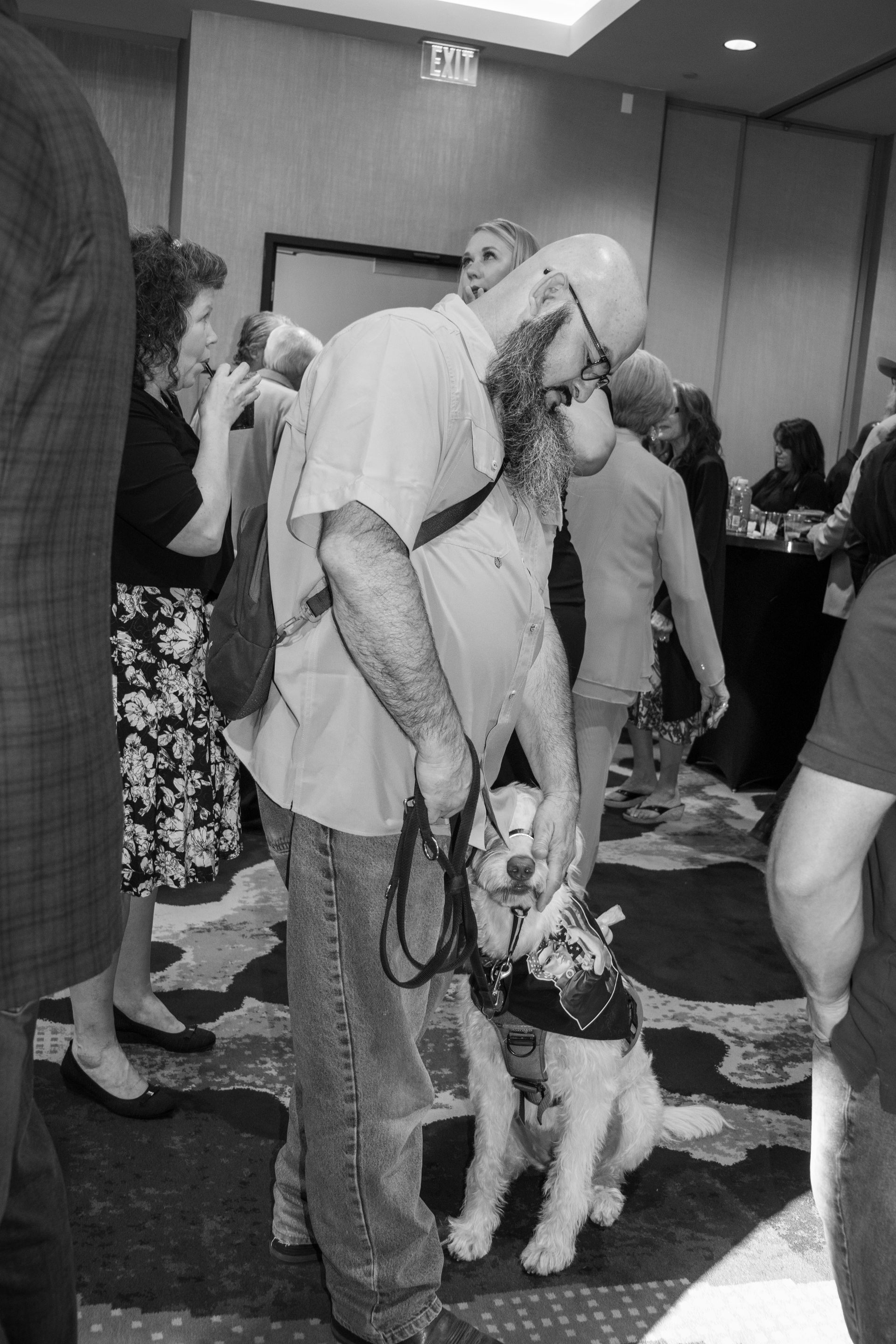 A person with a beard stands in a room, gently touching the face of a service dog wearing a harness.