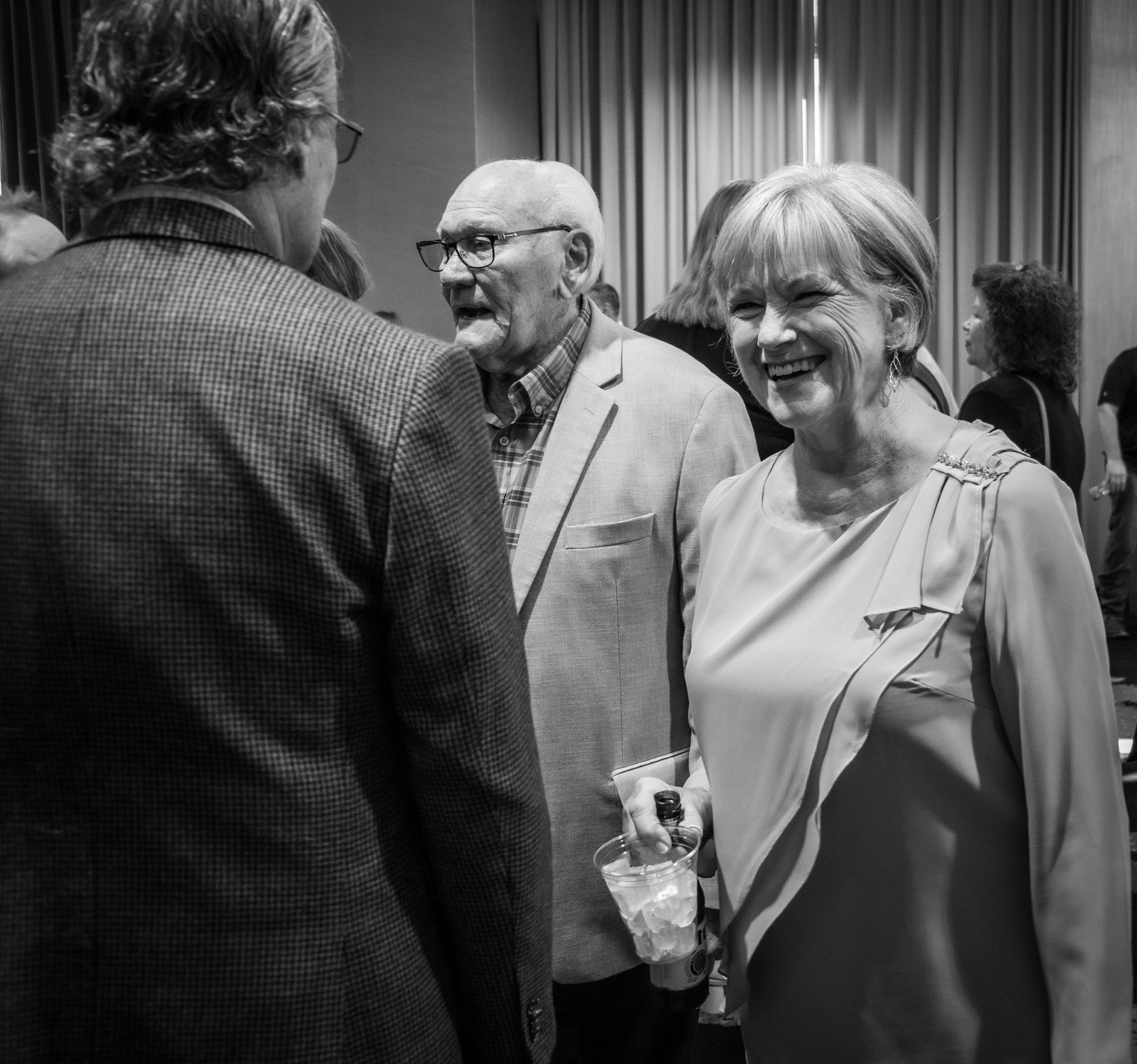 A group of people in formal attire stand in a room with curtained walls, smiling and conversing during an event.