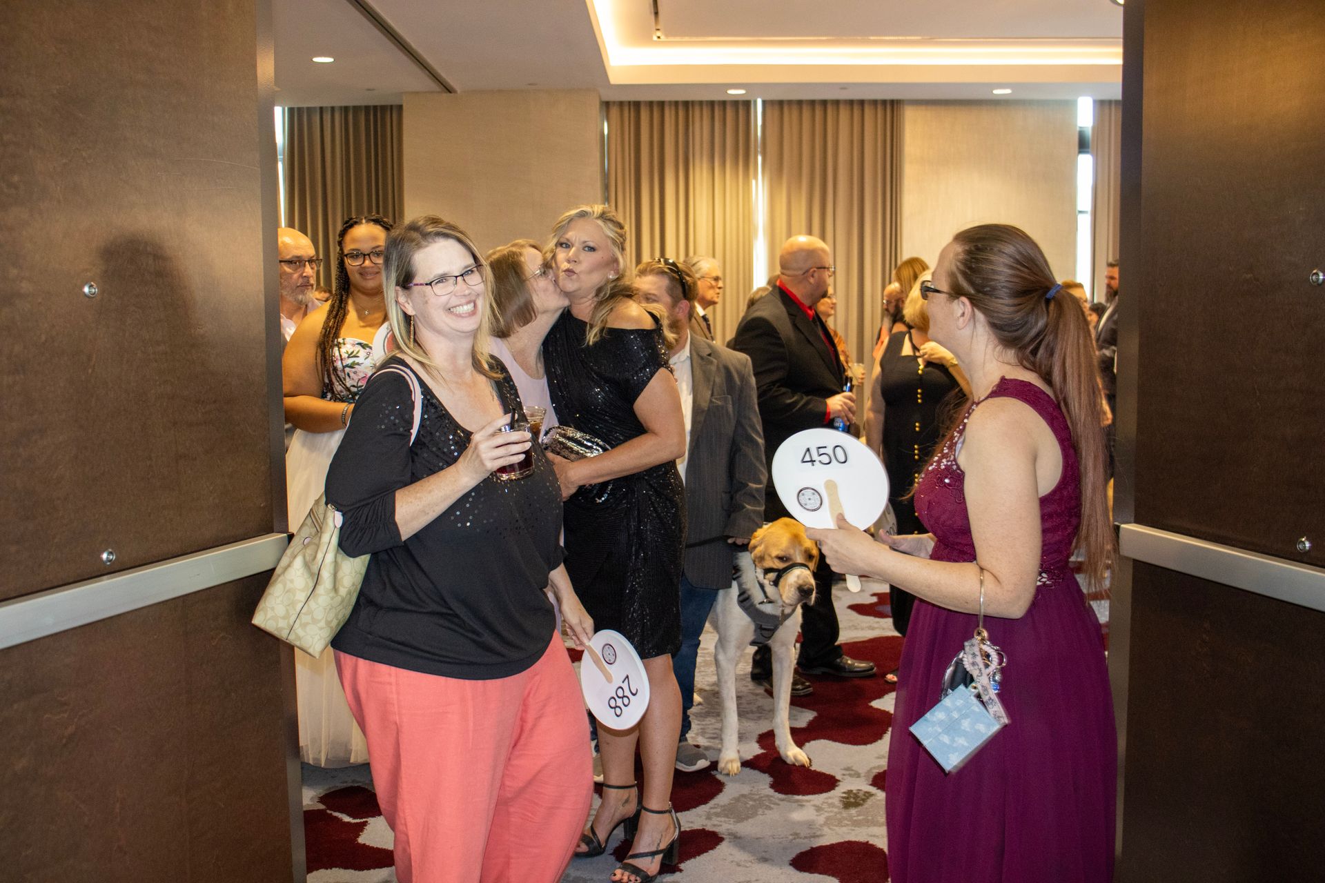 Attendees mingle in a hotel event space, with a dog present and individuals holding event programs and drinks.