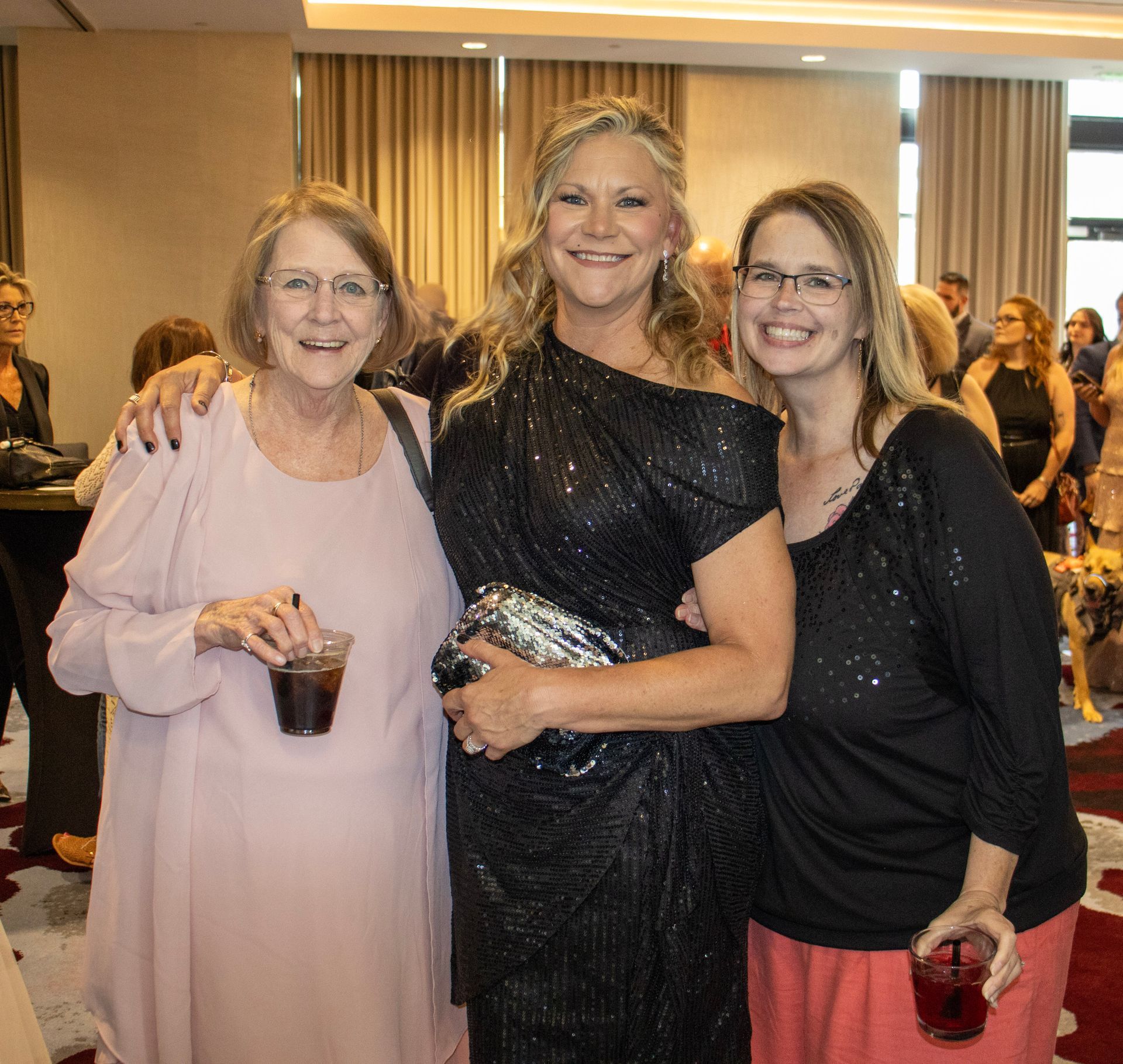 Three people pose for a photo at an indoor event, all smiling while holding glasses of dark liquid in a ballroom setting.