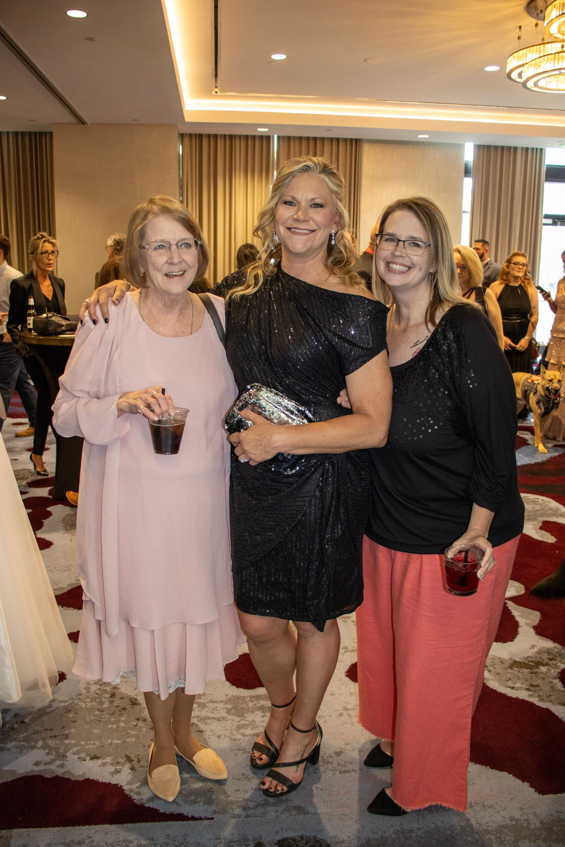 Three people pose together at an indoor event, smiling and holding drinks in a room with a patterned rug and chandeliers.