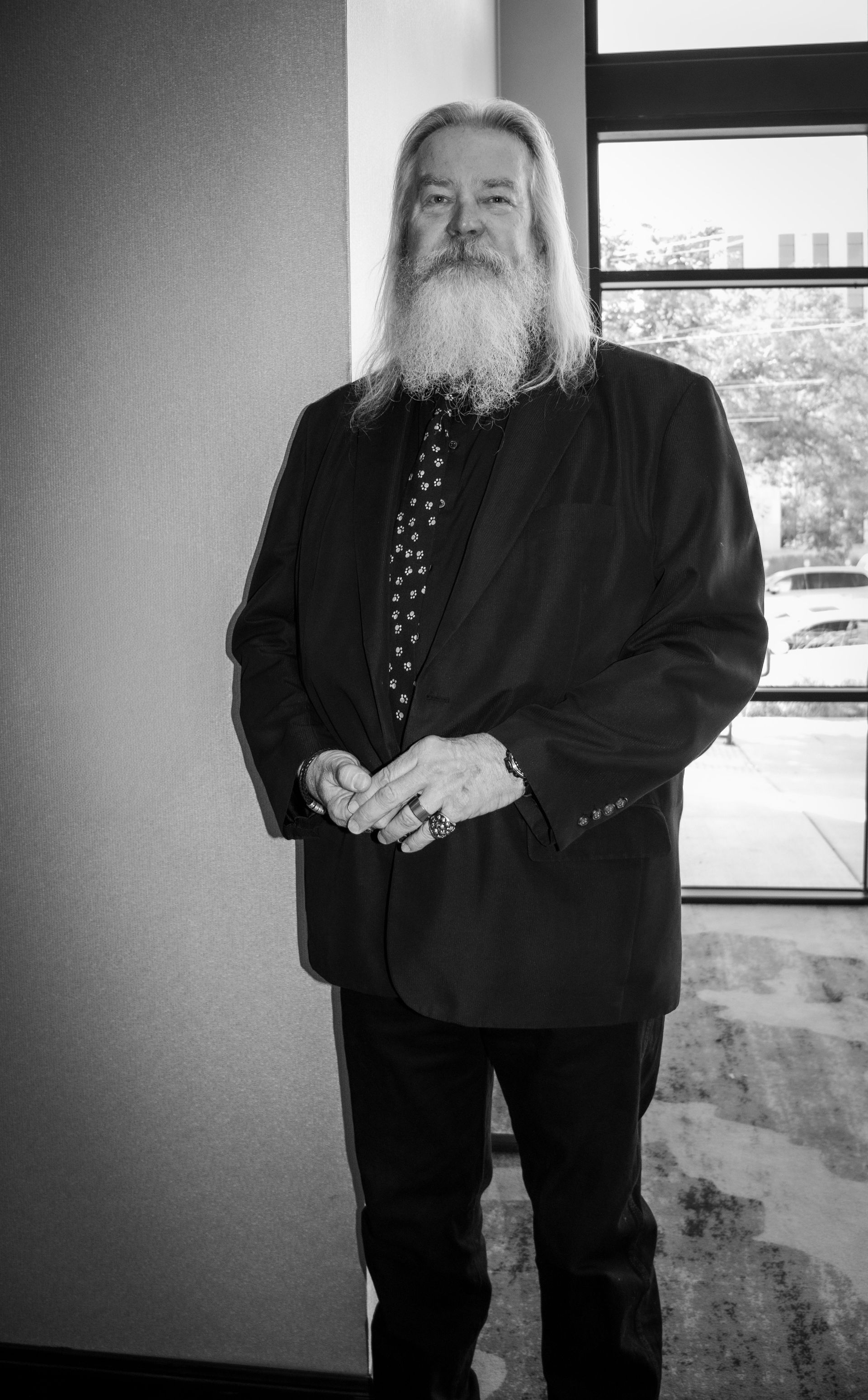 A person with a long beard, wearing a suit and patterned tie, stands with hands clasped in a brightly lit indoor space.