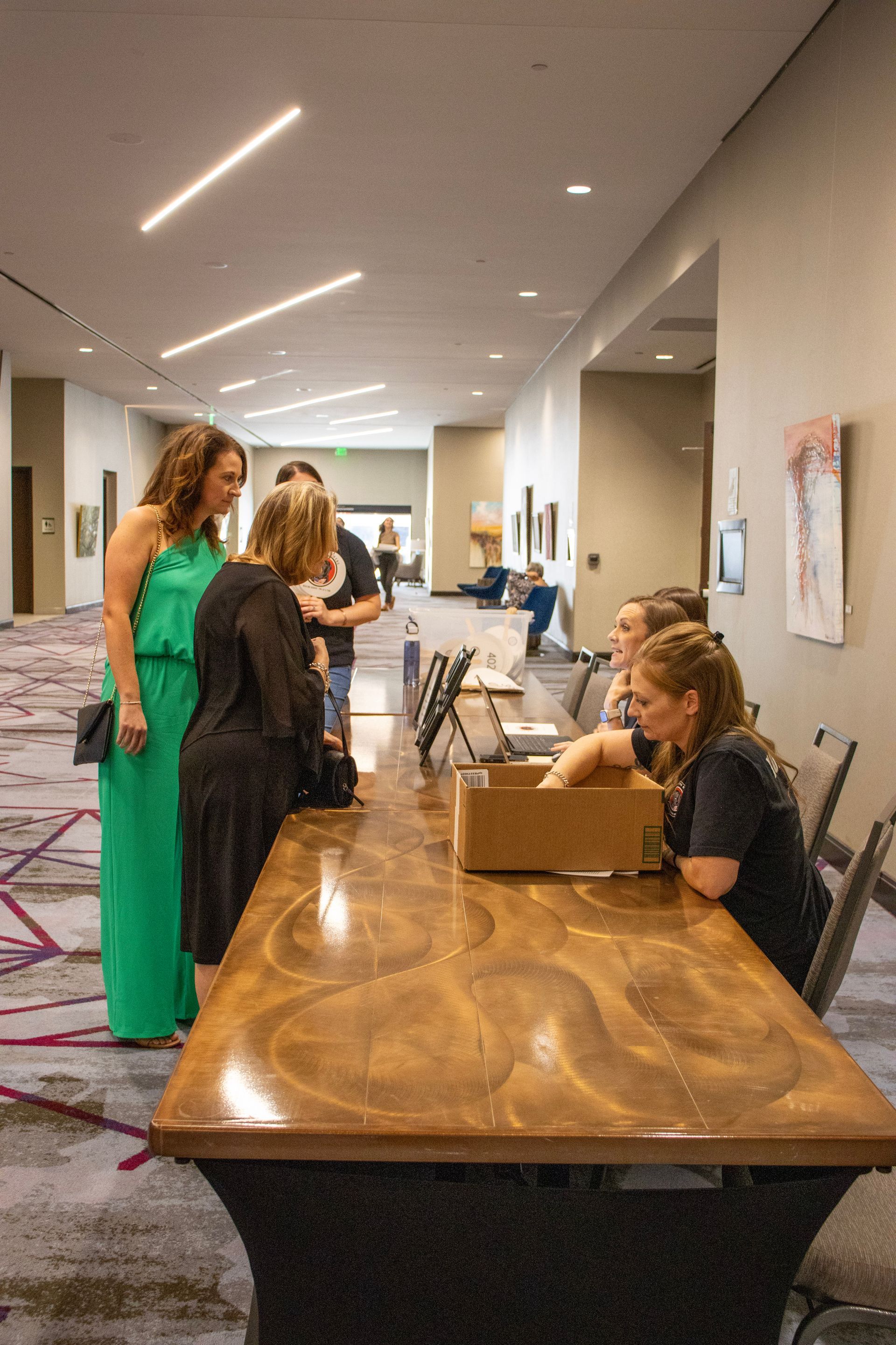 People stand at a long wooden registration table in a brightly lit hallway, interacting with a staff member.