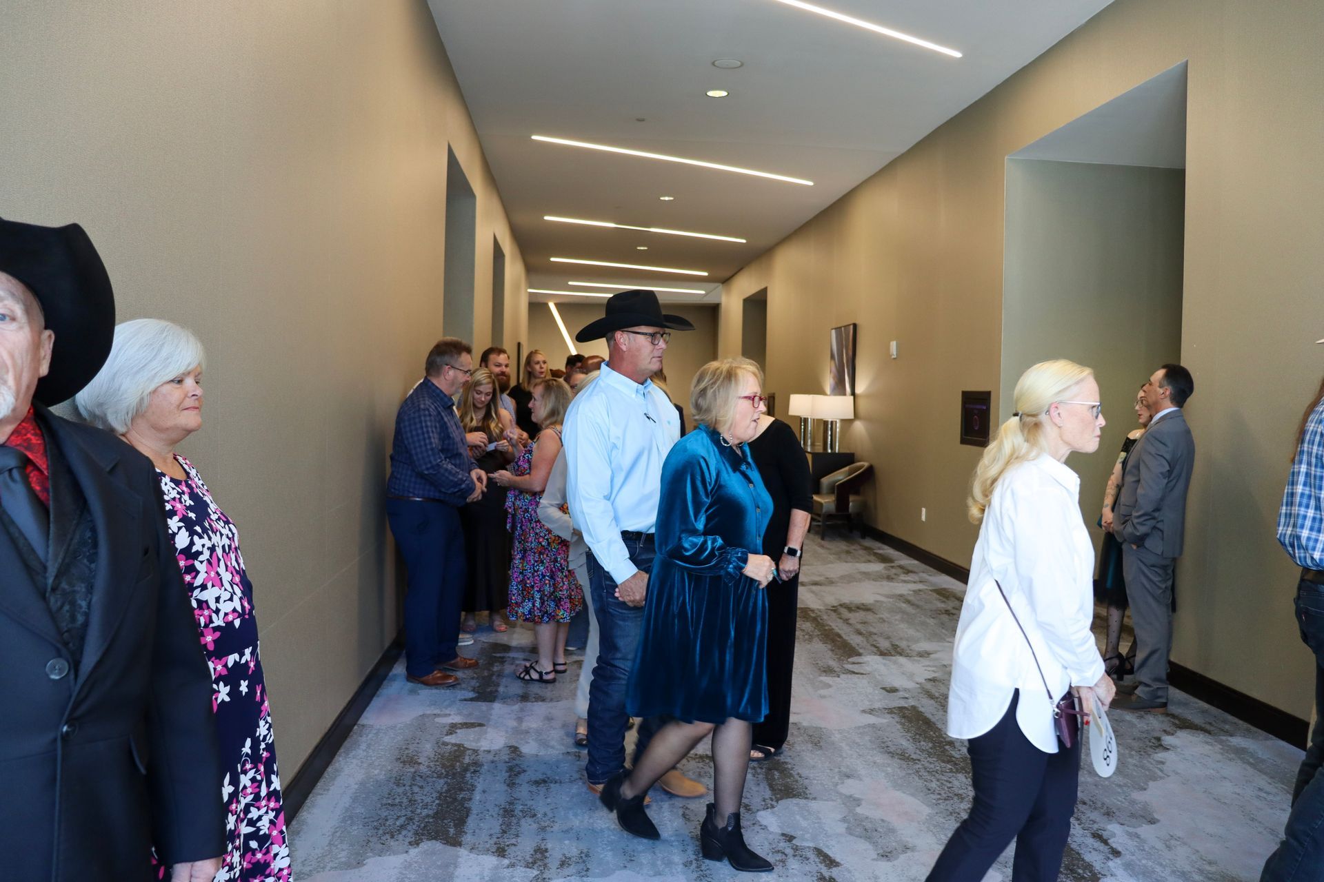 People stand in a line along a modern hallway with tan walls and patterned carpet, moving toward an event entrance.