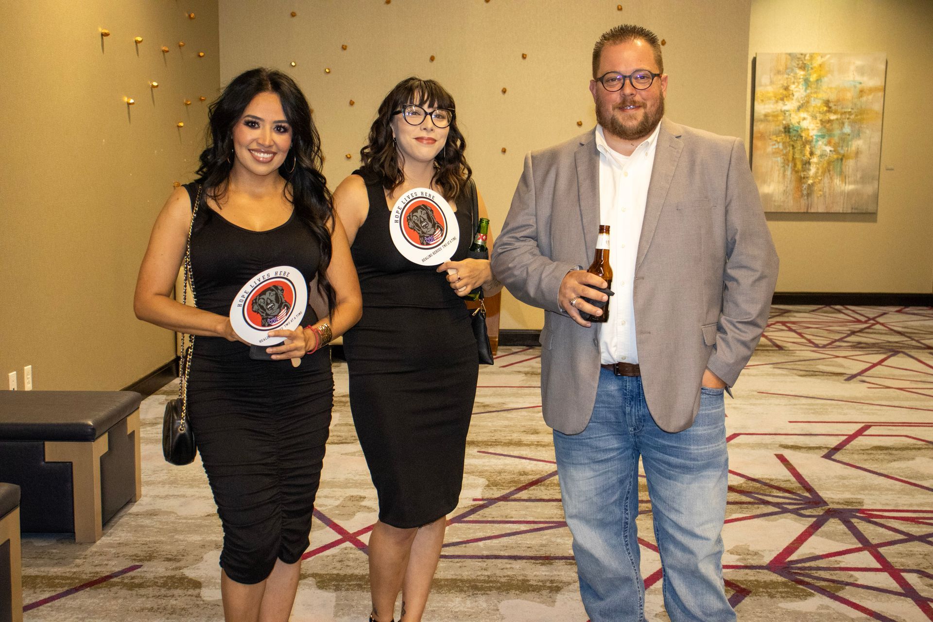 Two people in black dresses holding circular event signs and one person in a blazer holding a beer stand in a lobby.