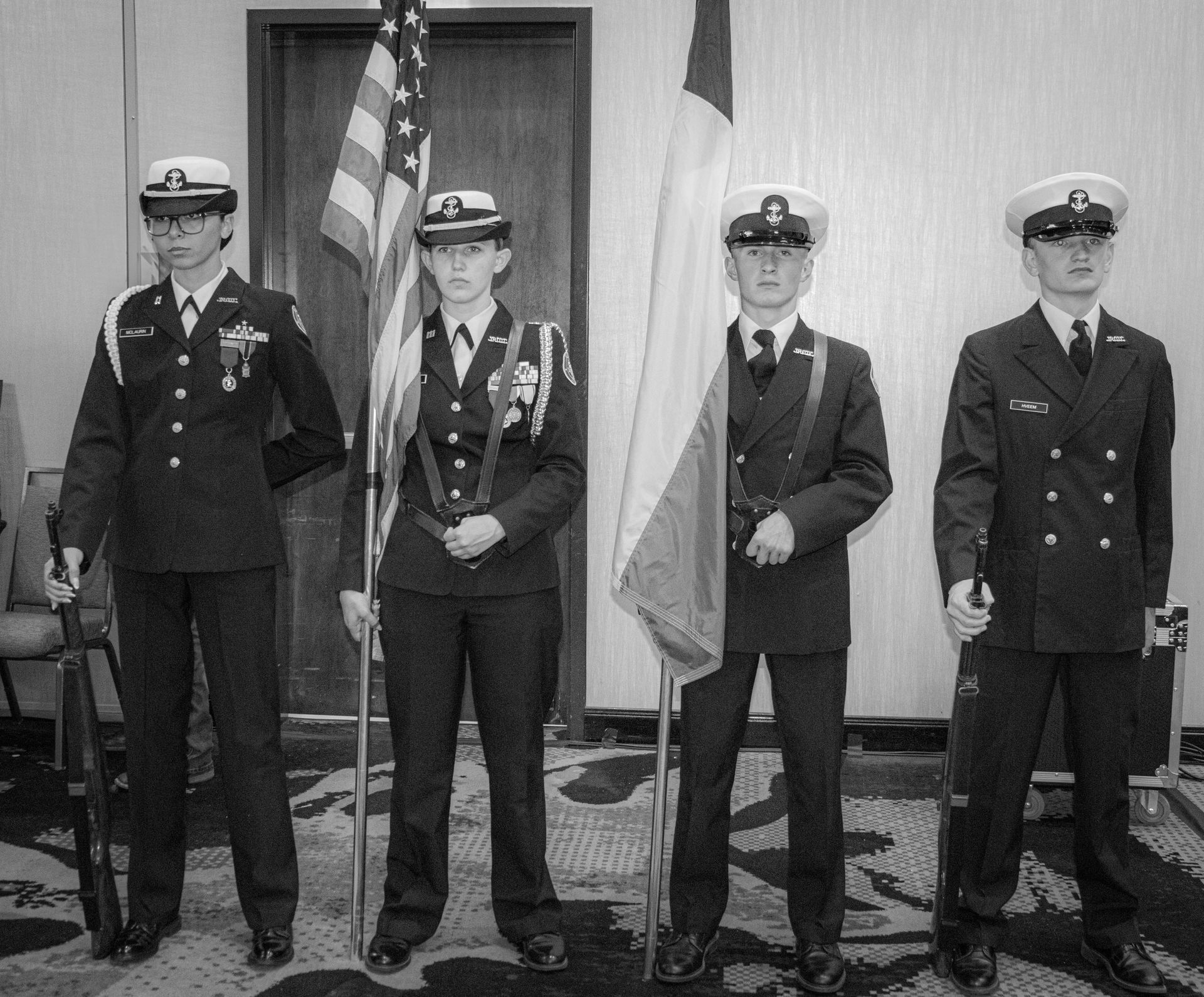 A four-person color guard in formal military dress uniforms stands in a line, holding two flags and two rifles.