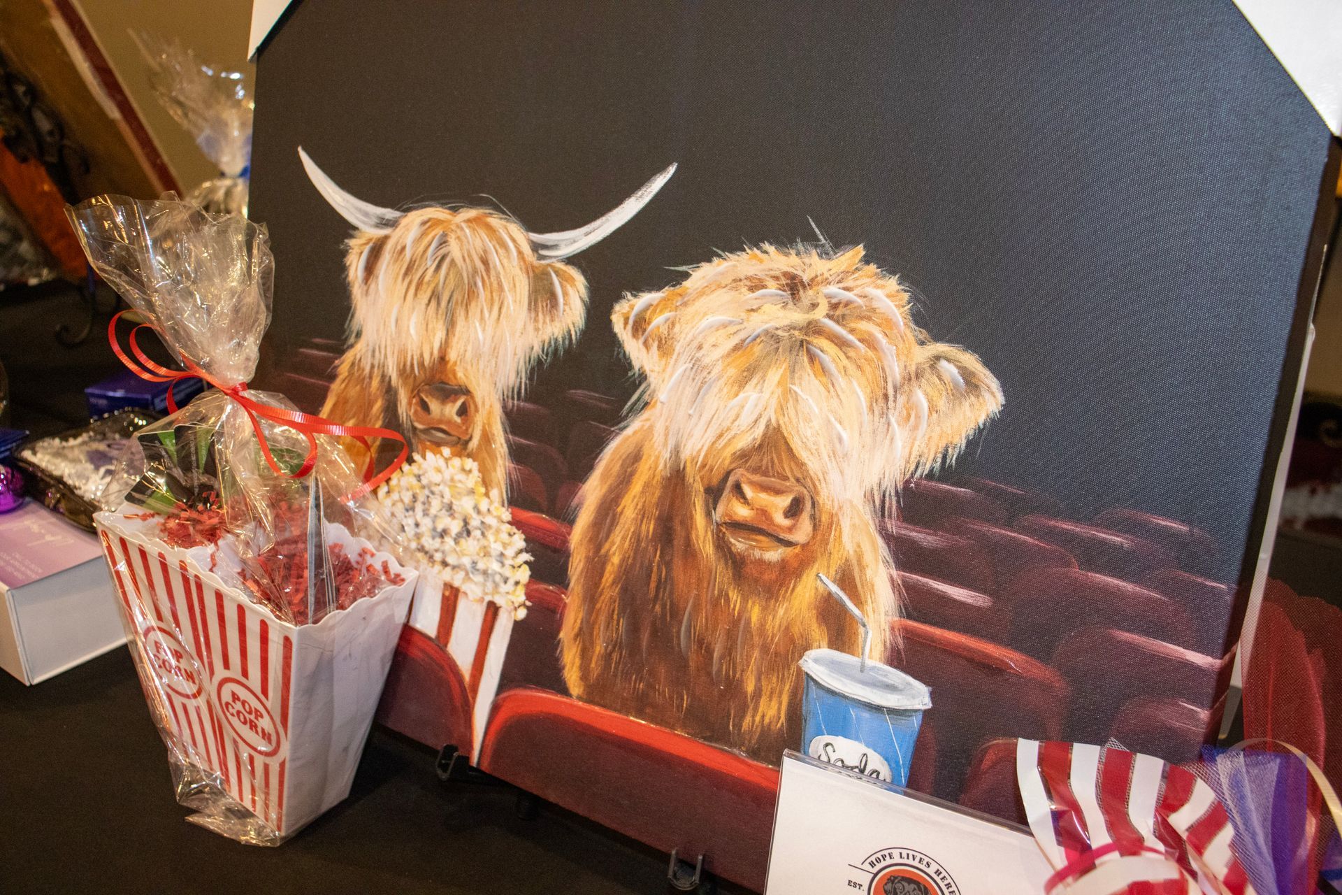 A painting of two Highland cows sitting in a movie theater with a popcorn bucket and a drink cup on a table in front.