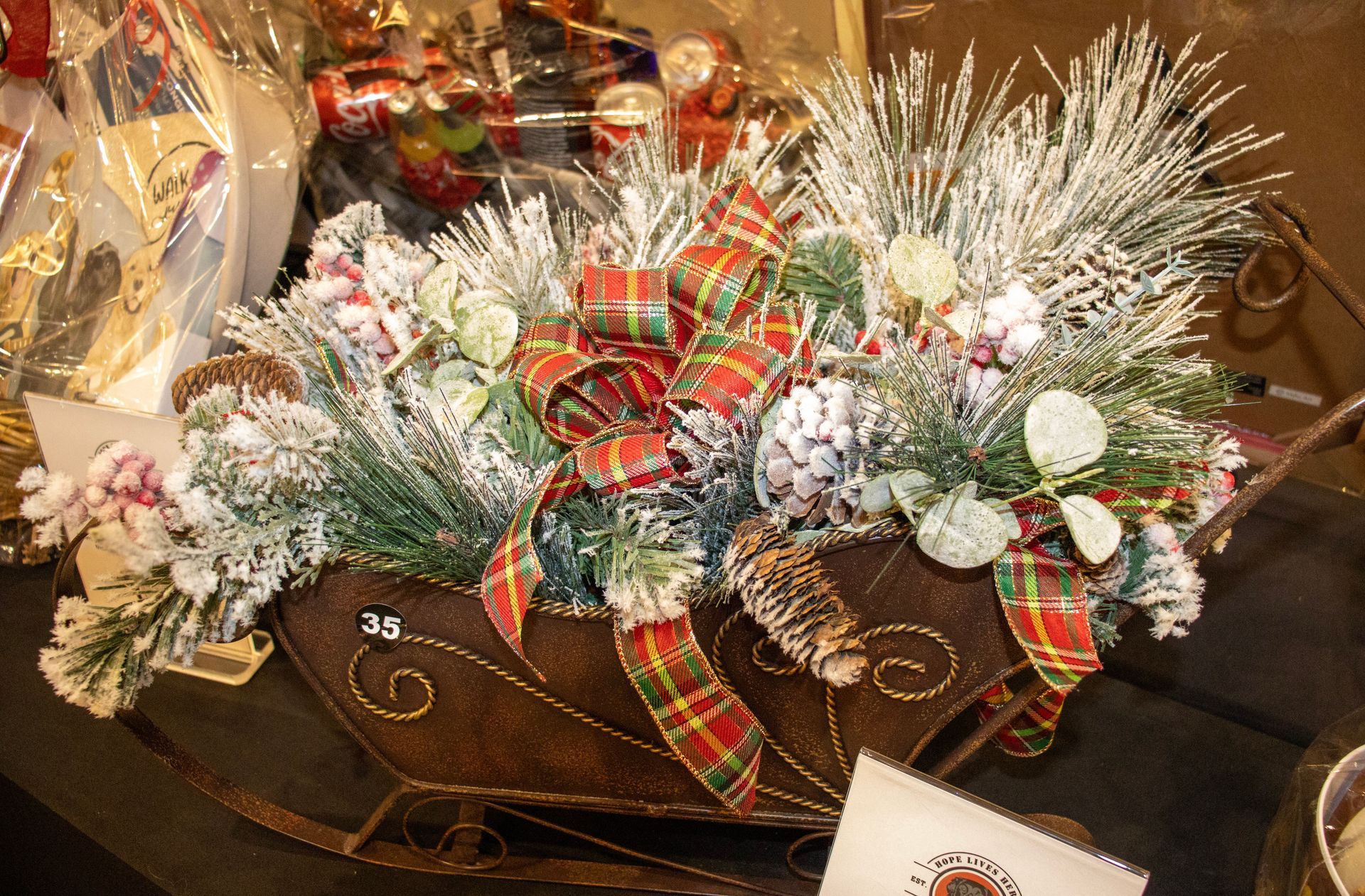 A decorative, rustic metal sleigh filled with snowy evergreen branches, pinecones, berries, and a plaid ribbon bow.