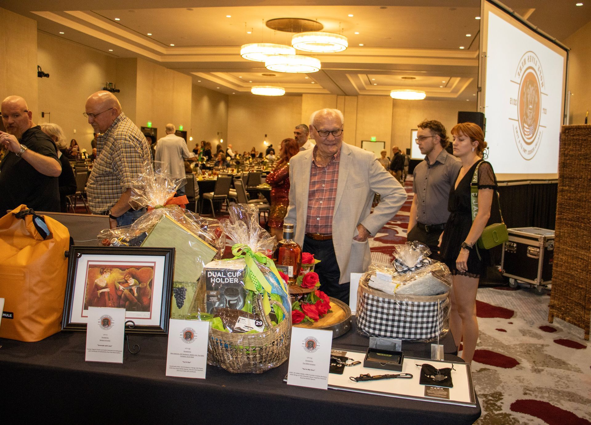 People browse auction baskets on a table at a brightly lit, large indoor banquet hall event.