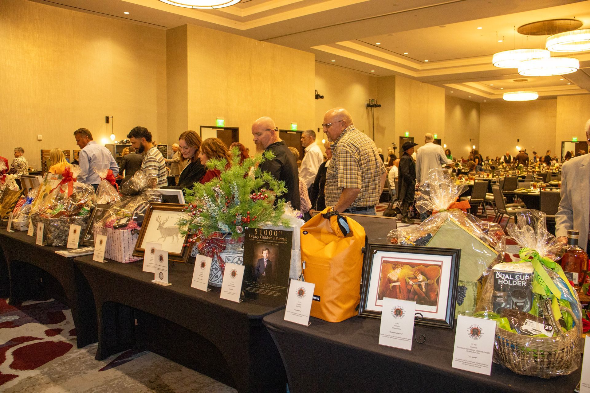 People browse a silent auction table filled with gift baskets and framed art in a large, well-lit banquet hall.
