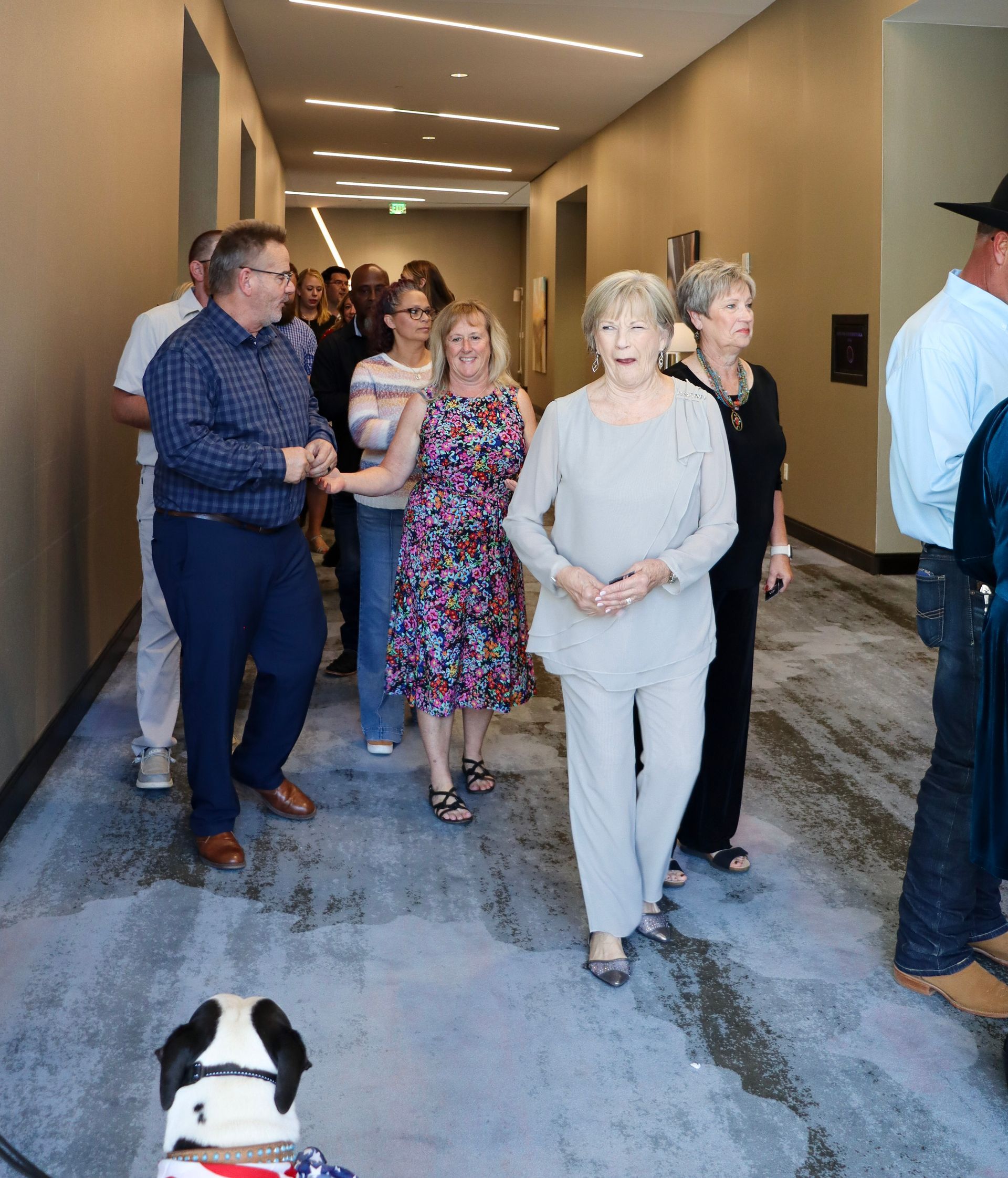 A group of people walk down a hotel hallway with patterned carpet, led by a small dog in the foreground.