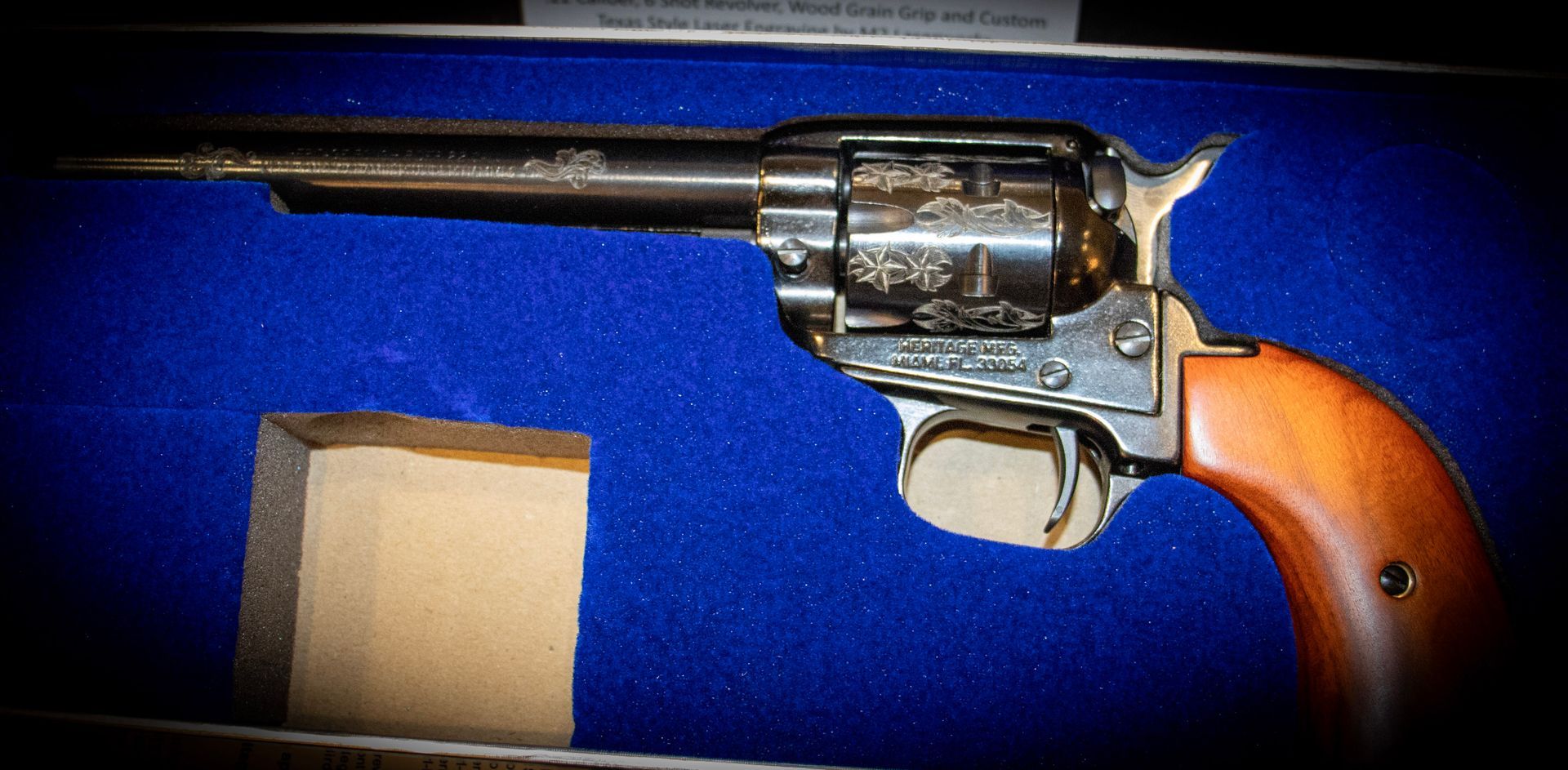 An engraved single-action revolver with a wooden grip, resting inside a display case lined with bright blue velvet.
