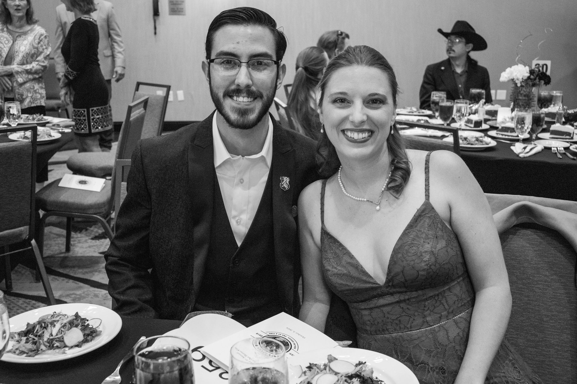 A man in a suit and a woman in a formal dress smiling at a table during a dinner event.