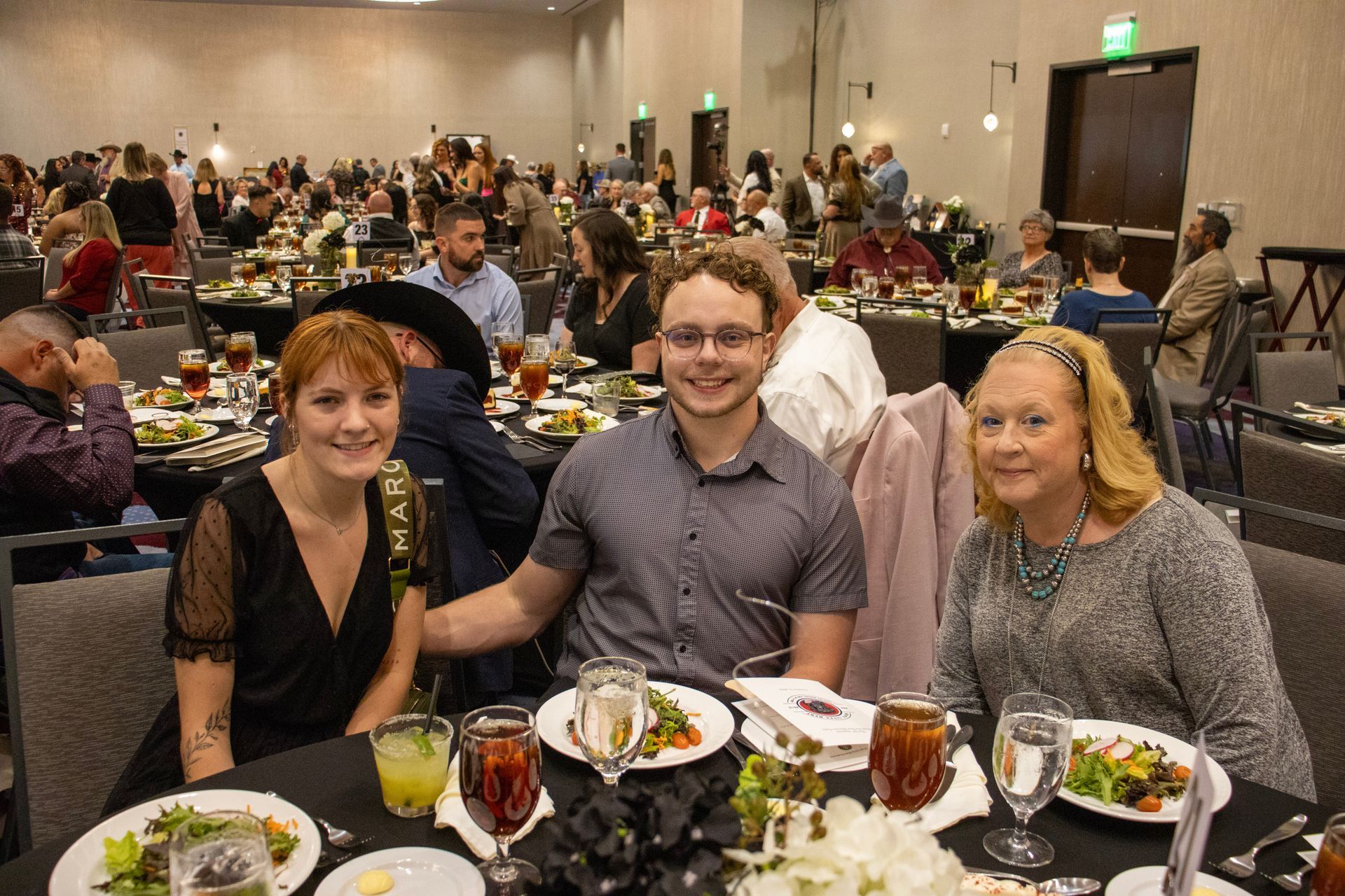 Three people seated at a table at a banquet, smiling for a photo with plates of food and drinks in front of them.