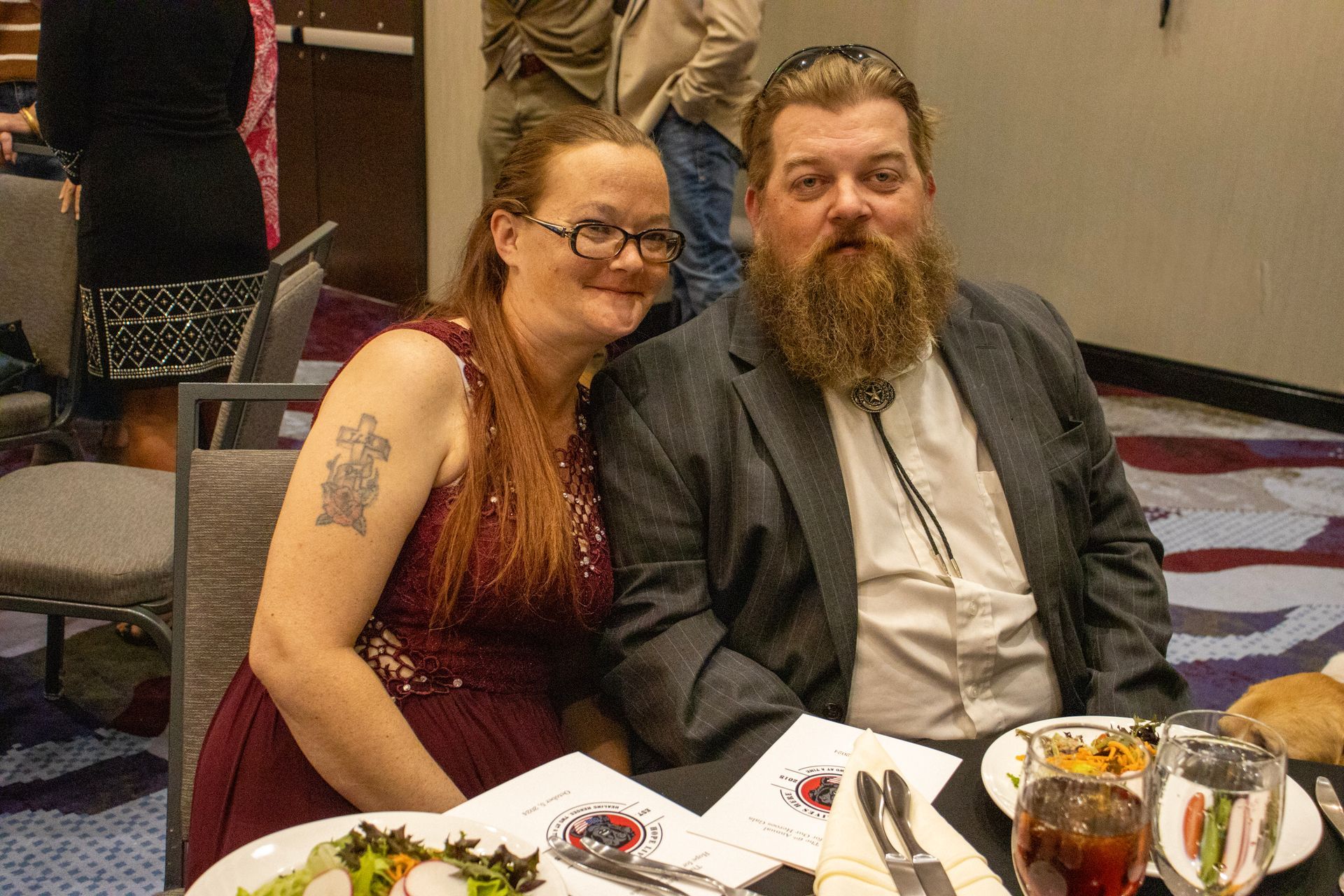 A person with glasses and a shoulder tattoo sits next to a bearded person wearing a blazer at a table with plates of food.