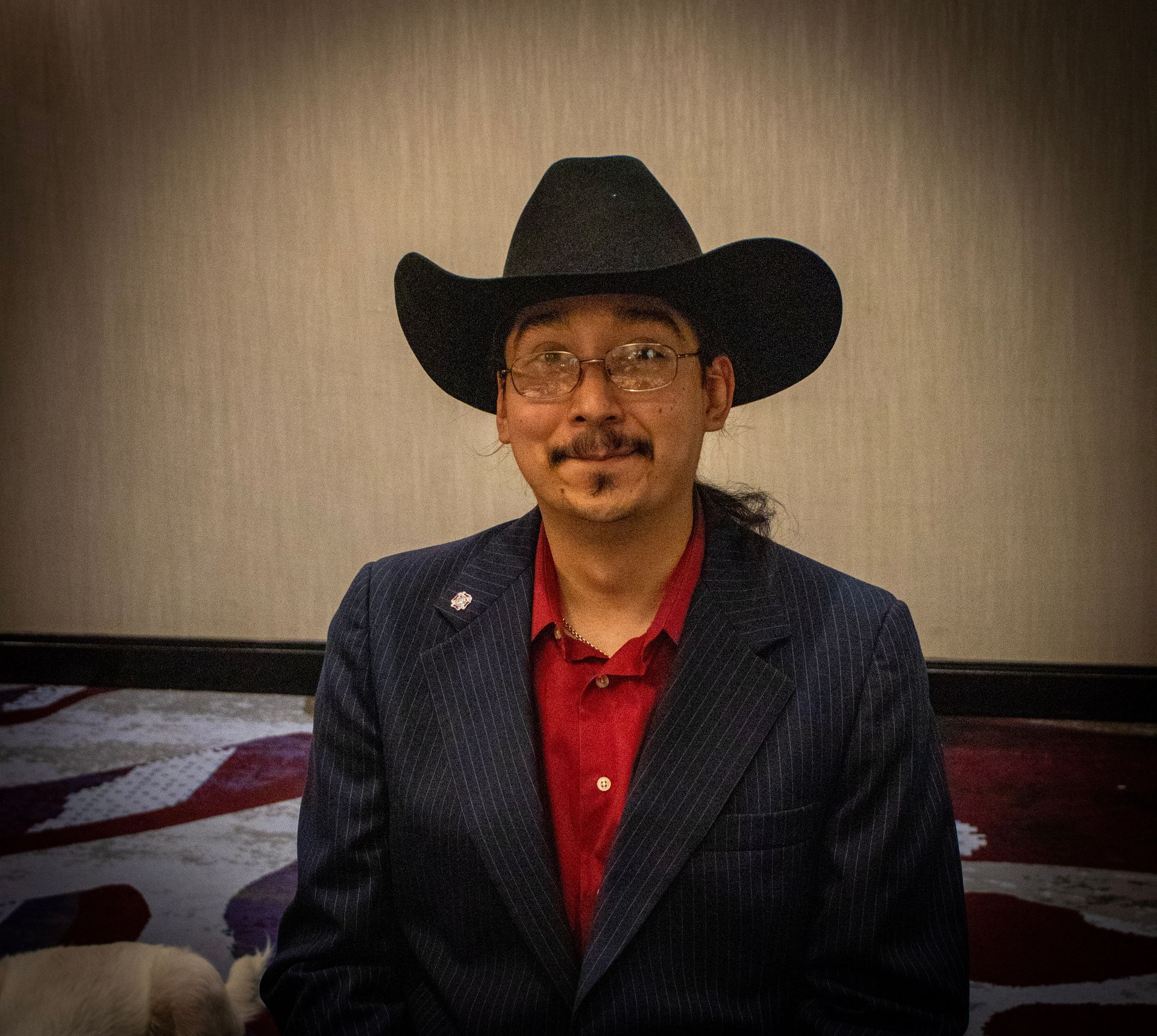 A person with glasses and a black cowboy hat wears a red shirt under a dark blue patterned blazer against a neutral wall.