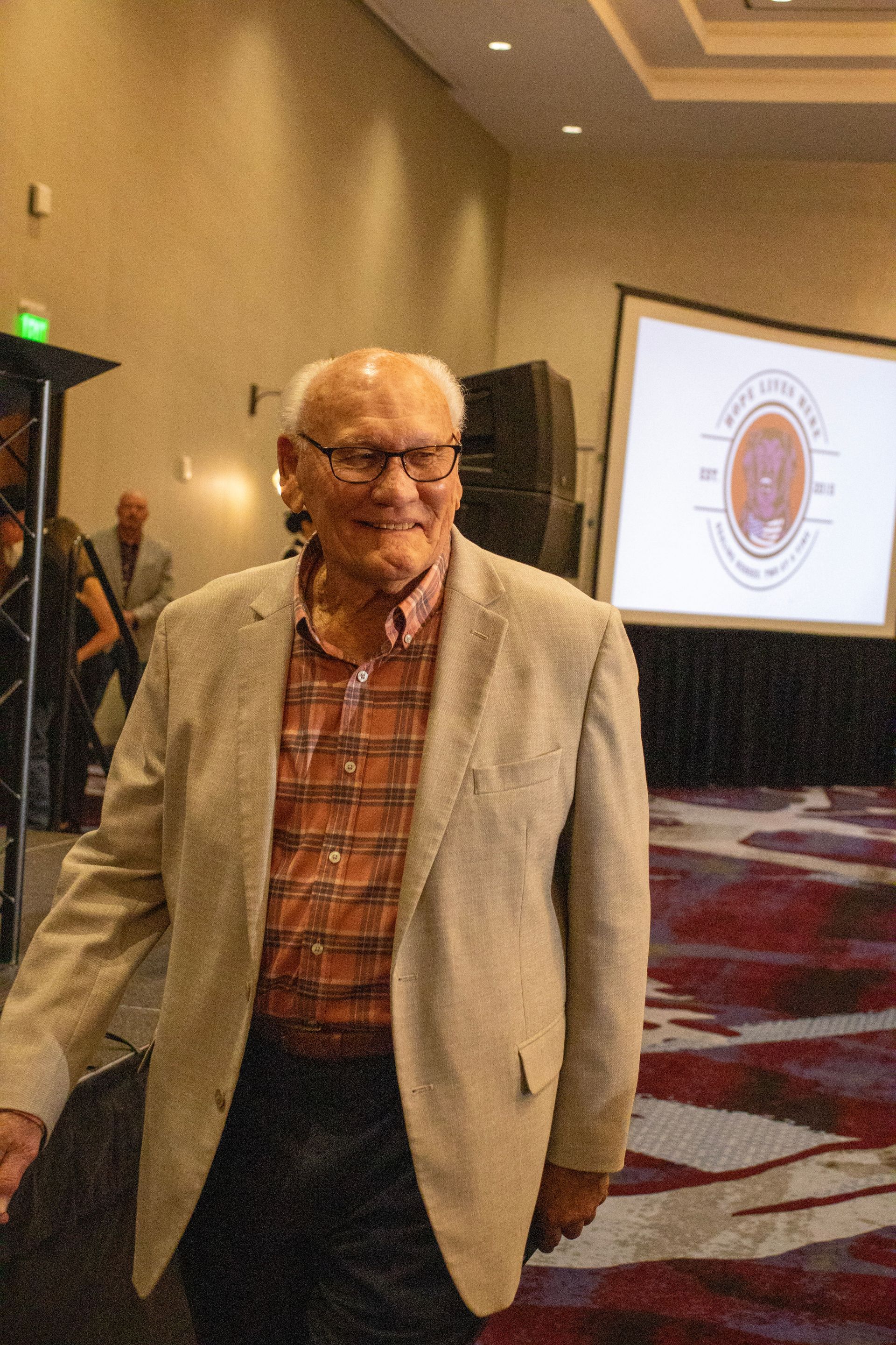 A smiling person in a tan blazer and plaid shirt walks through a room with a projector screen showing a circular logo.